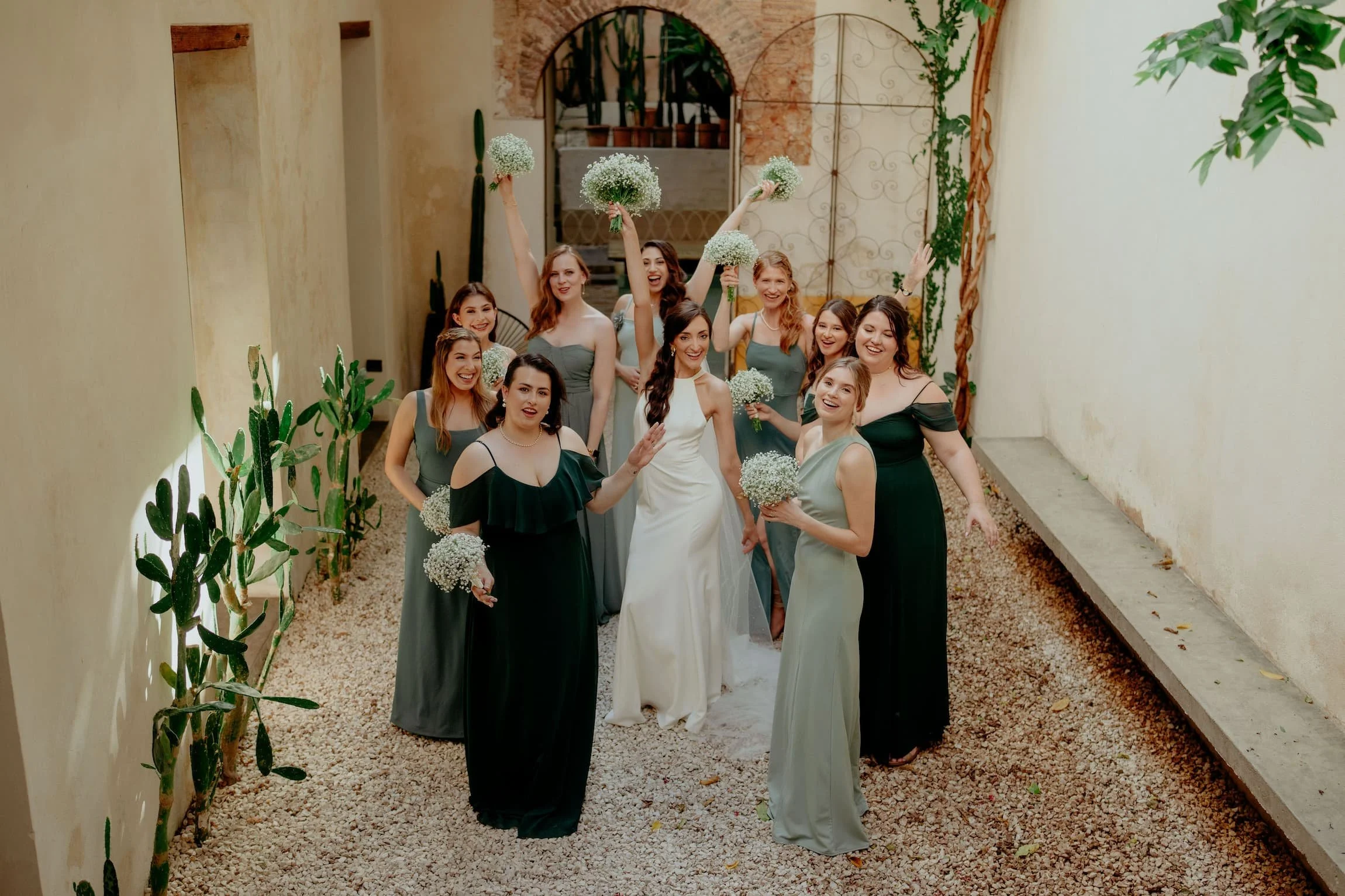 A group of nine women dressed in wedding attire, holding small bouquets of white flowers, celebrating in a narrow outdoor corridor with beige walls, gravel ground, and green plants.