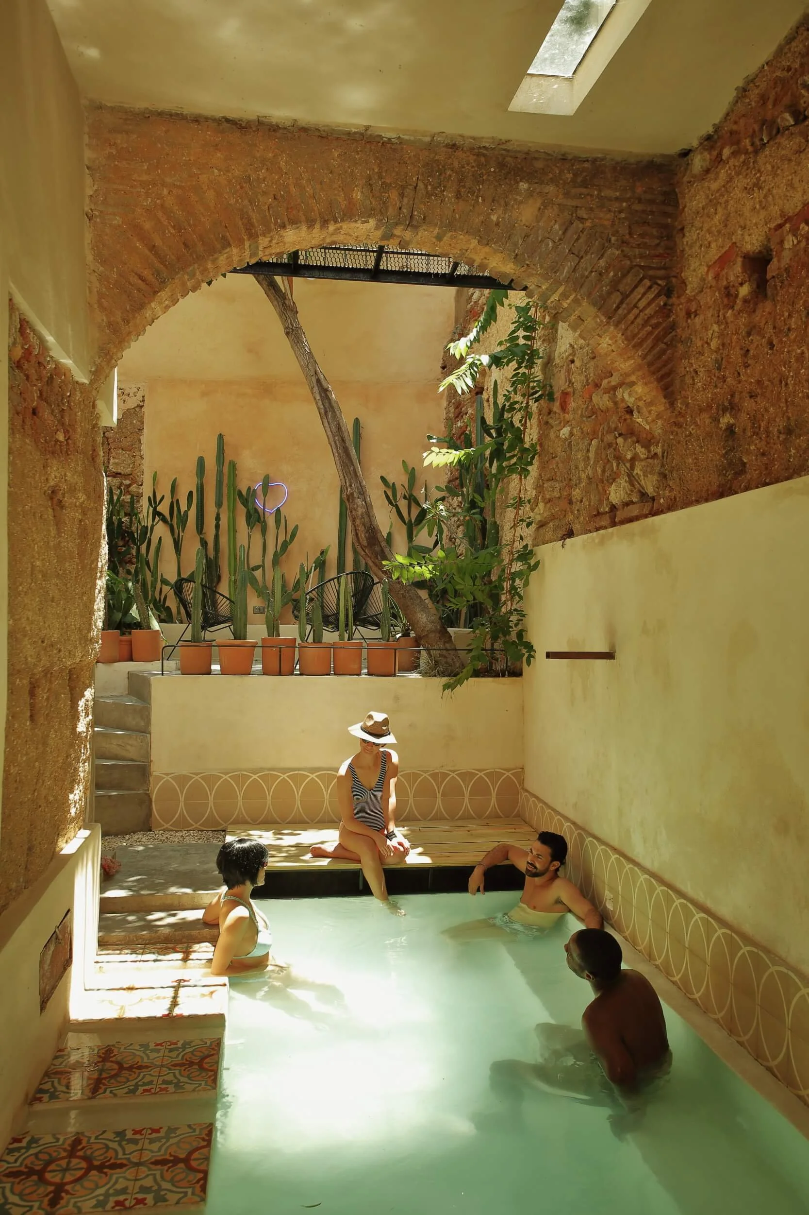 People relaxing in an indoor pool, with potted cacti and plants on a raised platform in the background, and a stone and brick arch overhead.