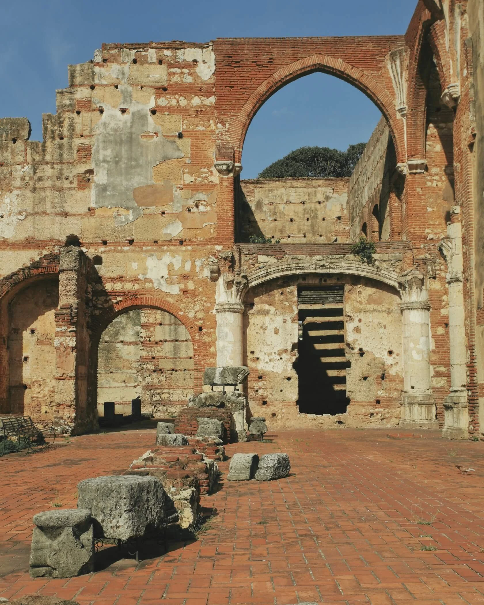 Ruins of an ancient building with red brick and stone walls, arched openings, and a staircase leading to an upper level, set against a blue sky.