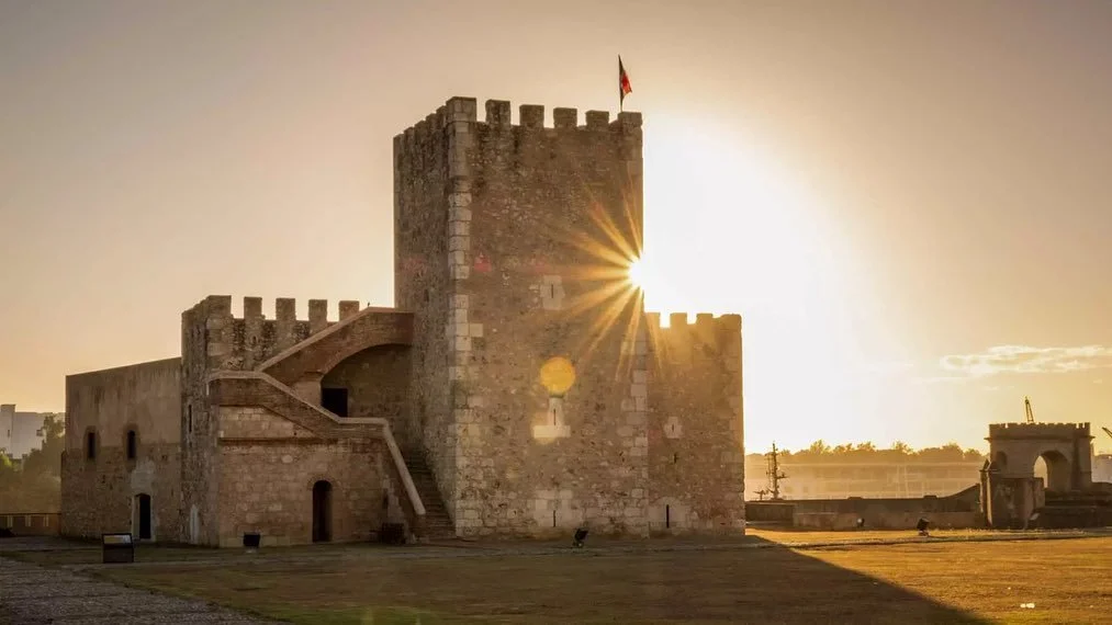 A historic stone castle with a flag on top, illuminated by the setting sun, with a clear sky and some trees in the distance.