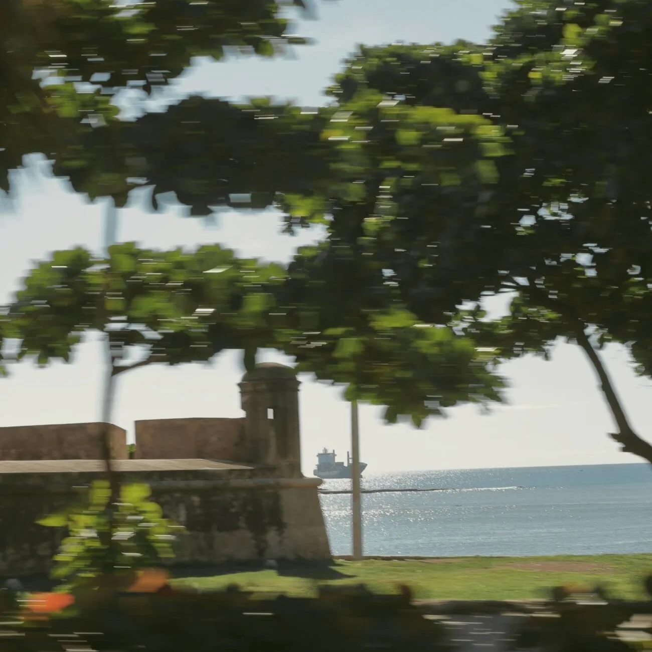 Blurred photo of a seaside with trees and a historic fort, with a ship in the water in the background.