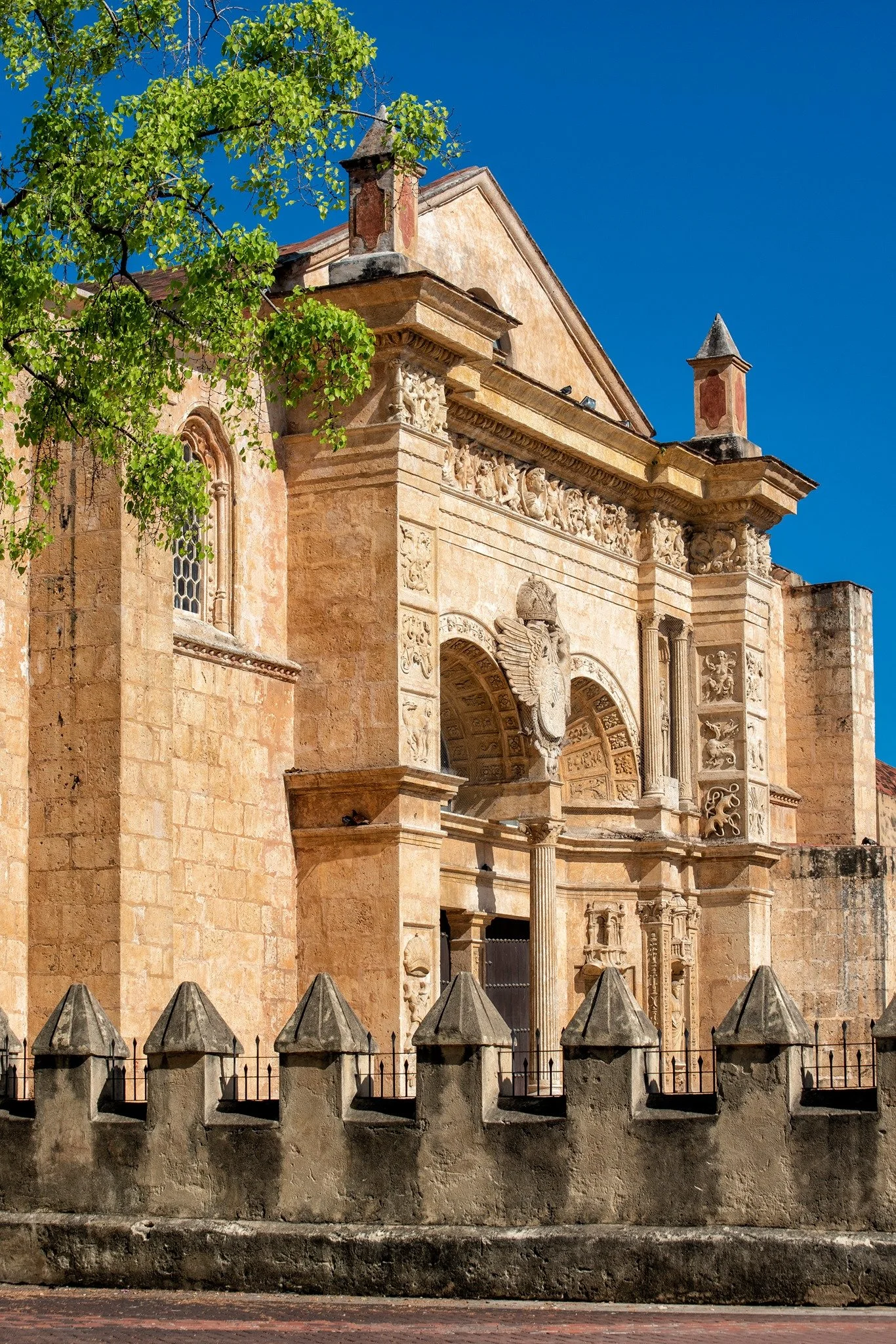 Historic stone building with ornate architectural details, topped with small pointed towers, surrounded by a stone fence and a tree with green leaves in front, under a clear blue sky.