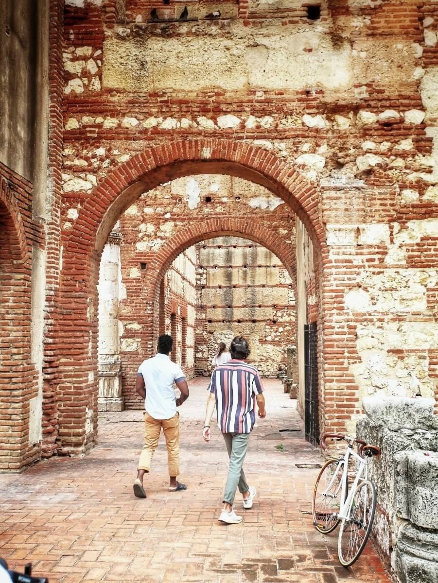 Three people walking through ancient brick arches in a historic site, with a bicycle leaning against a stone wall on the right.