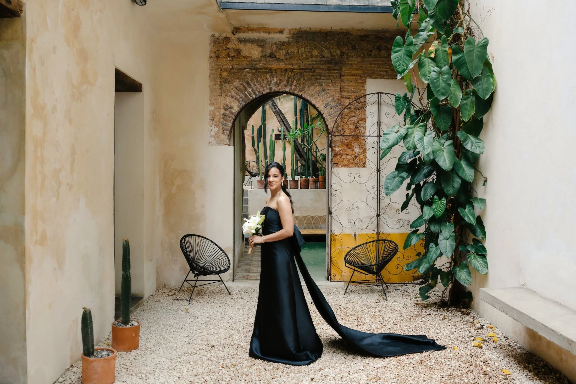 A woman in a strapless black dress holding a bouquet of white flowers standing in a courtyard with potted cactus, large green leaves, exposed brick archway, and black metal chairs.