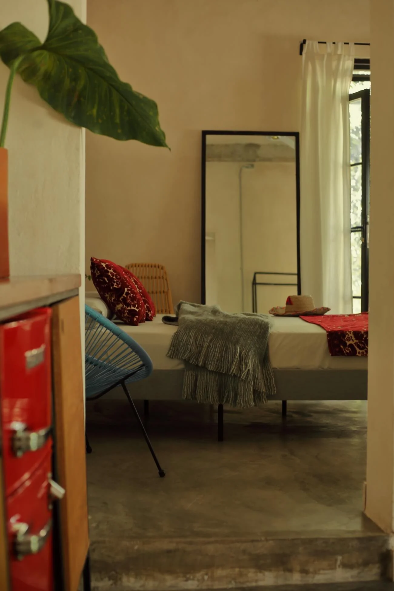 Interior of a cozy, well-lit bedroom with a bed, patterned pillows, a blanket, a hat, curtains, a mirror, a chair, and a plant in the corner.