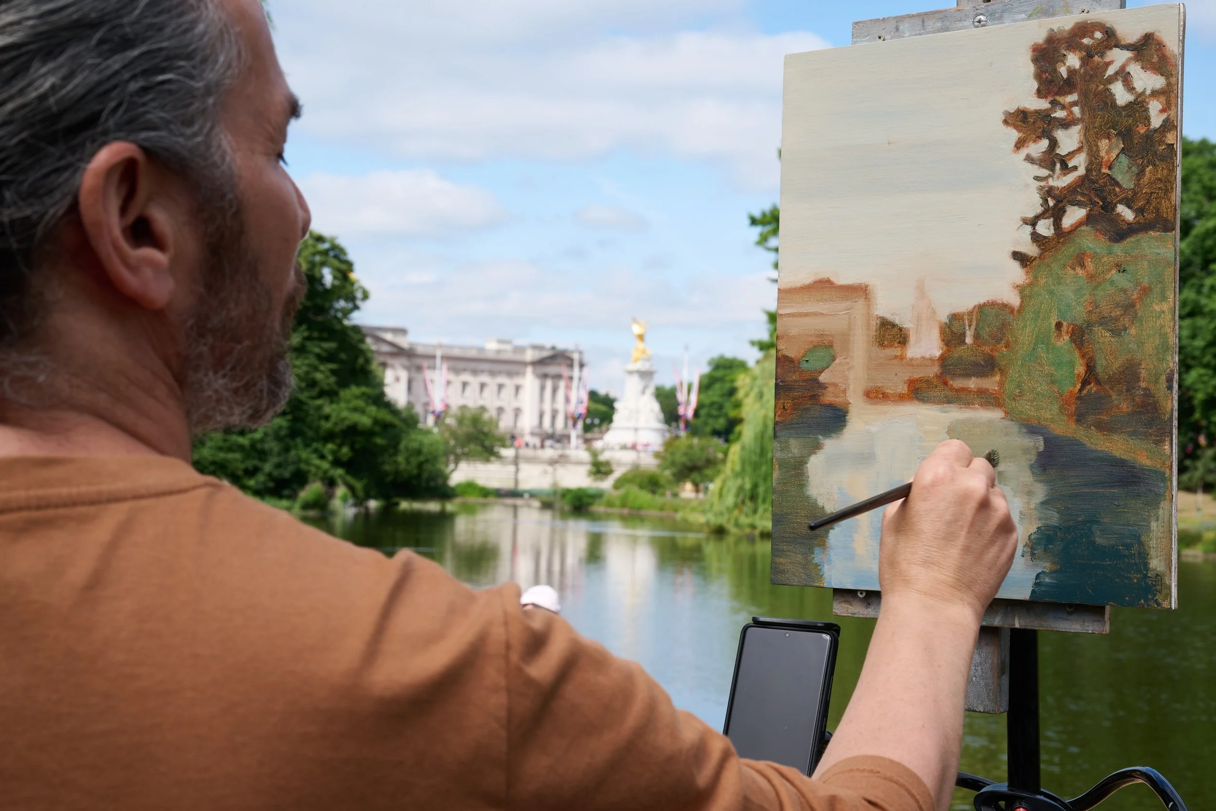 Man painting a landscape on an easel outdoors near a river, with a large building and a statue in the background.