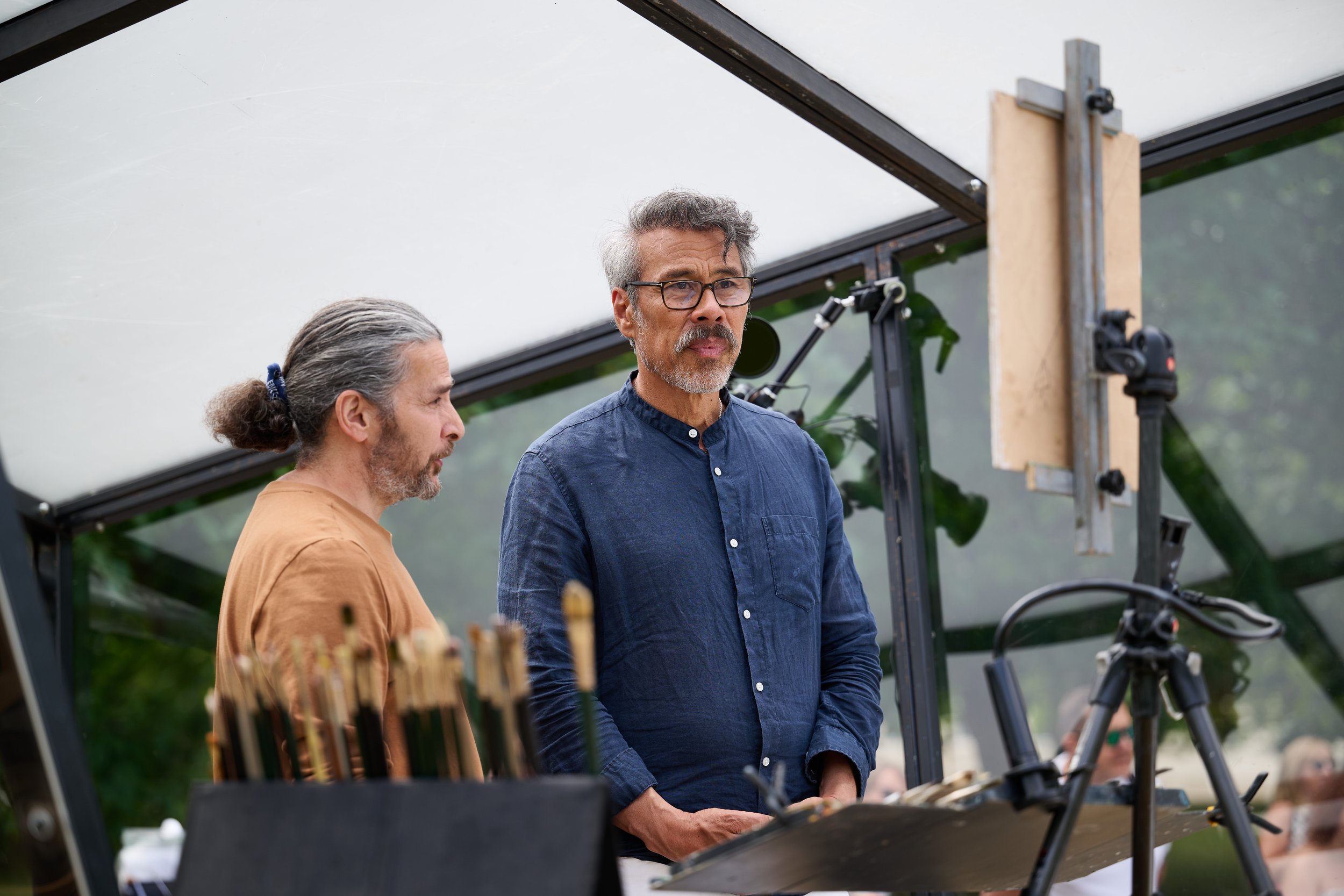 Two men standing outdoors near a solar panel, with one man wearing a blue shirt and glasses and the other man in a brown shirt and gray hair tied back, looking at something out of frame, possibly during a discussion or presentation.