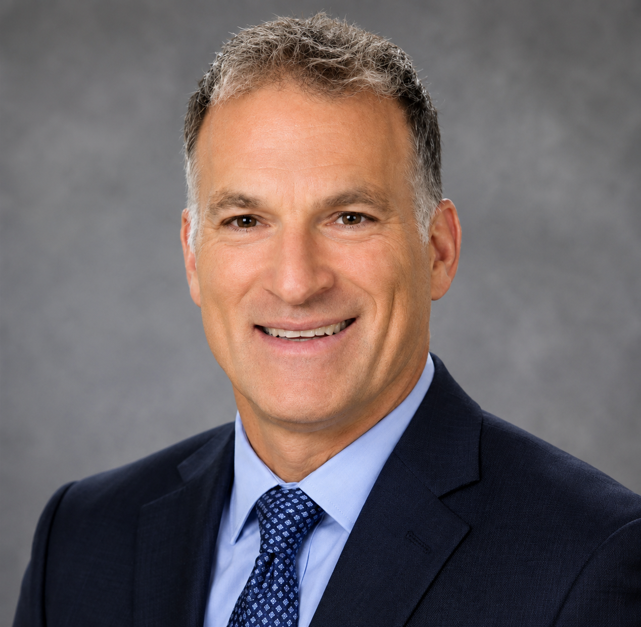 Headshot of a middle-aged man in a dark suit, light blue dress shirt, and patterned tie, smiling against a gray background.