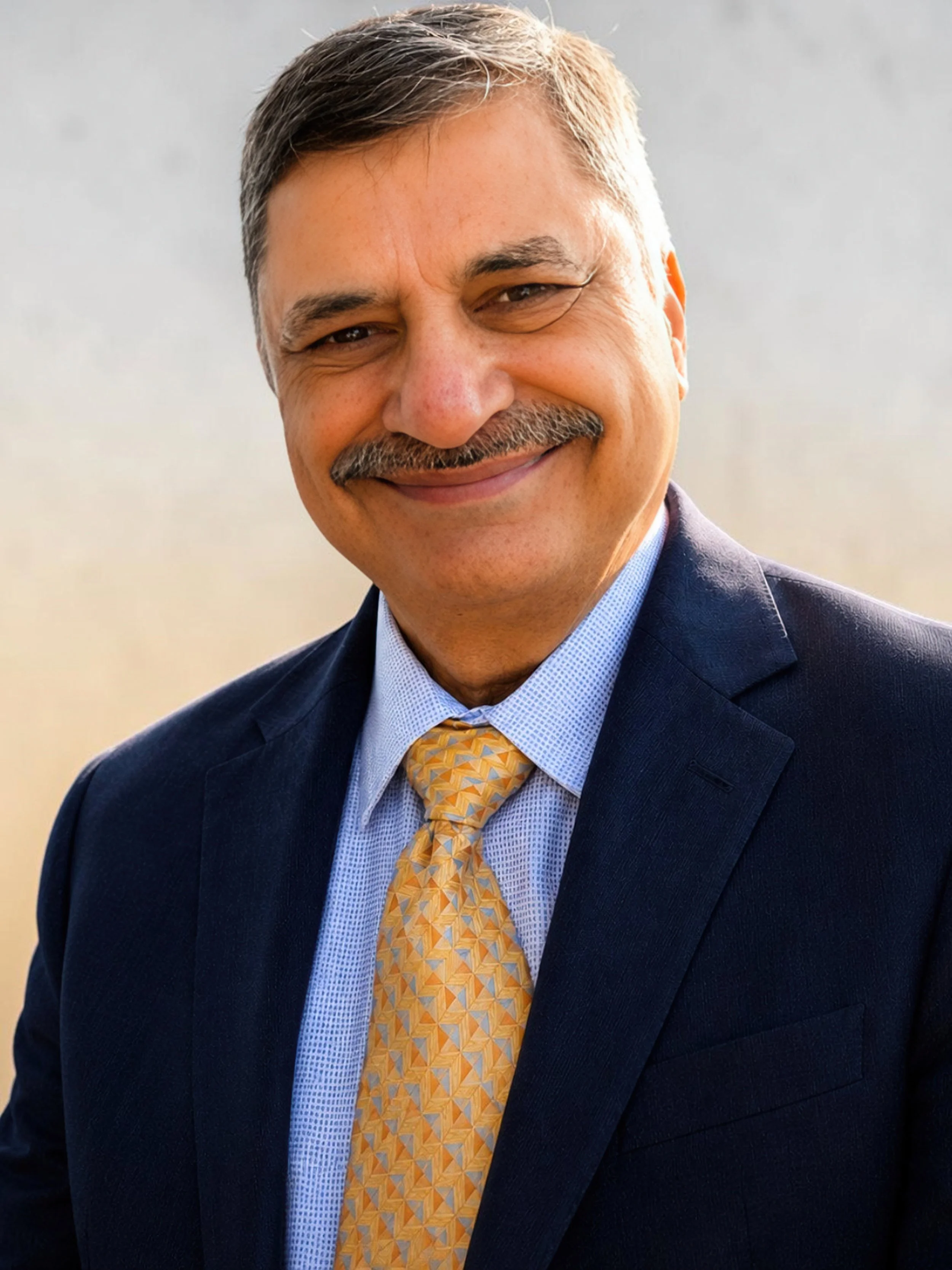 A middle-aged man with gray hair and a mustache dressed in a navy blue suit, light blue shirt, and patterned yellow tie, smiling.