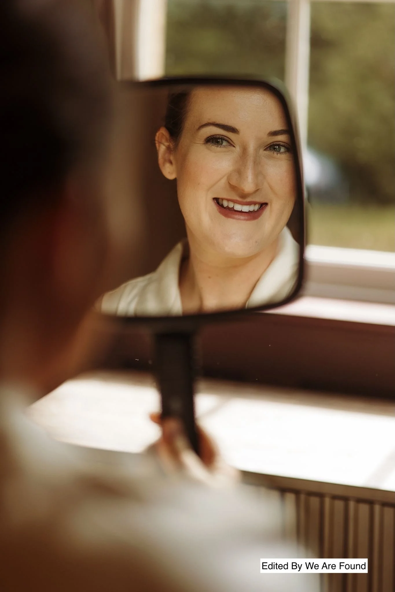A woman with light skin, dark hair, and blue eyes is smiling and looking into a mirror, with a blurred background outside the window.