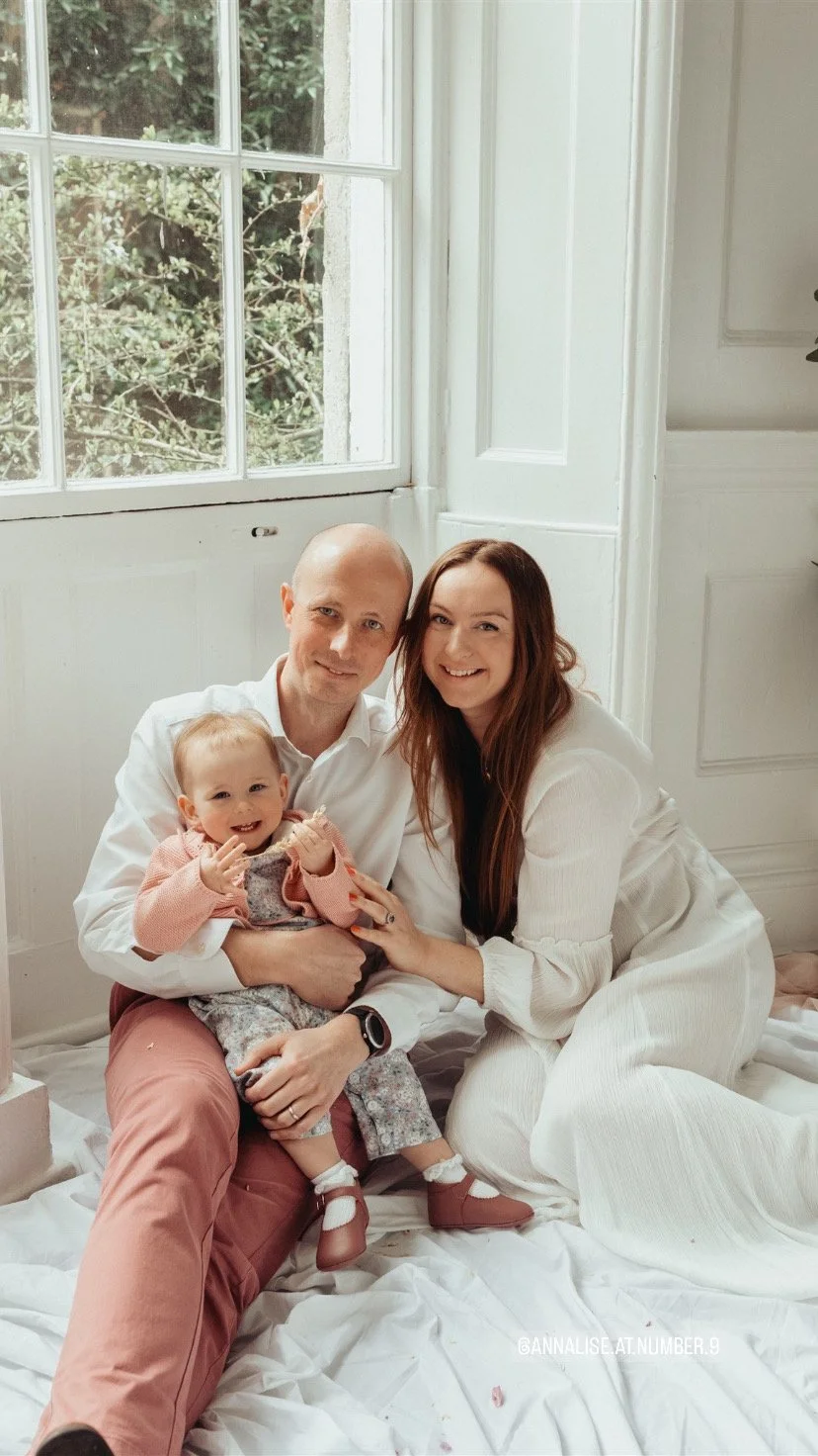 A family of three sitting on a white floor by a large window. A man is holding a toddler in his lap, and a woman is leaning close to them, all smiling.