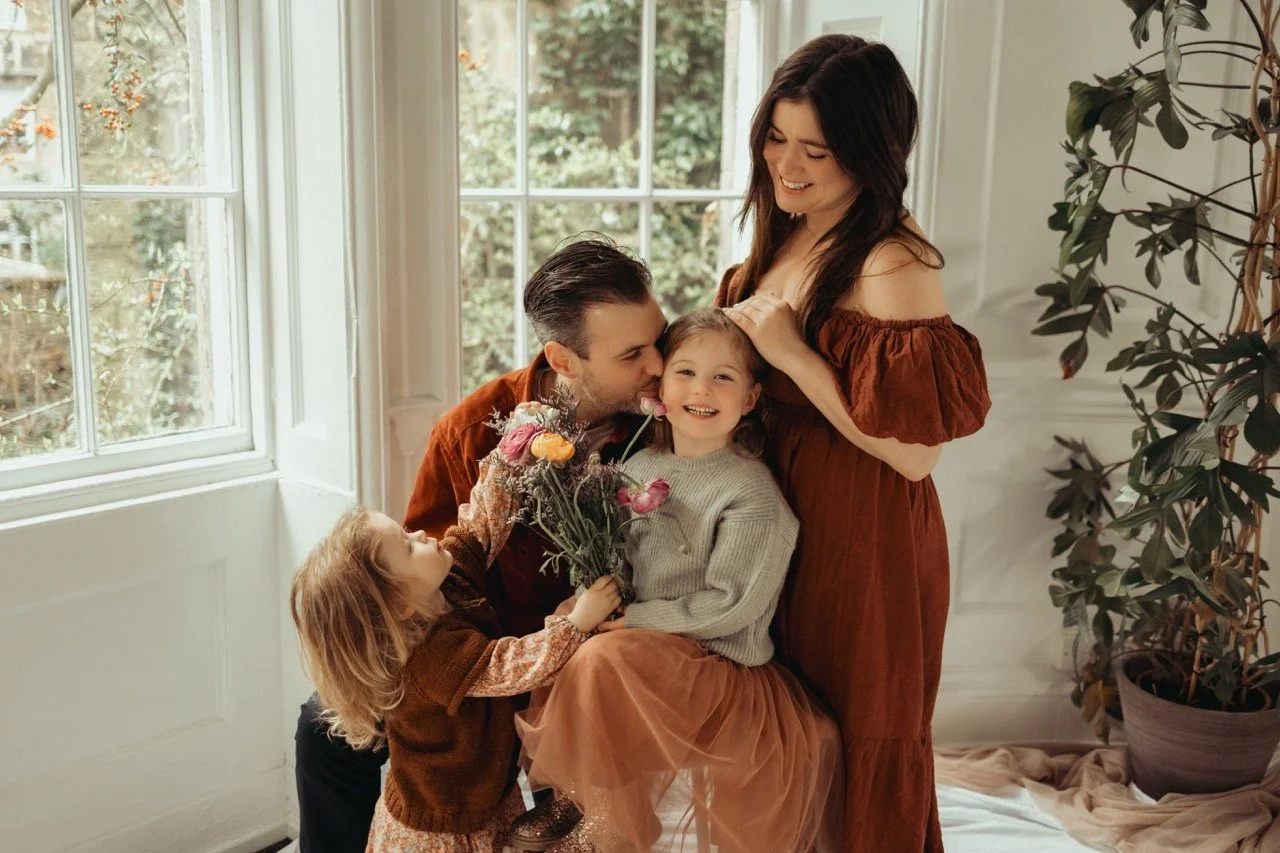 A family of four smiling indoors by a window, a man, woman, and two children holding flowers, dressed in autumn colors.