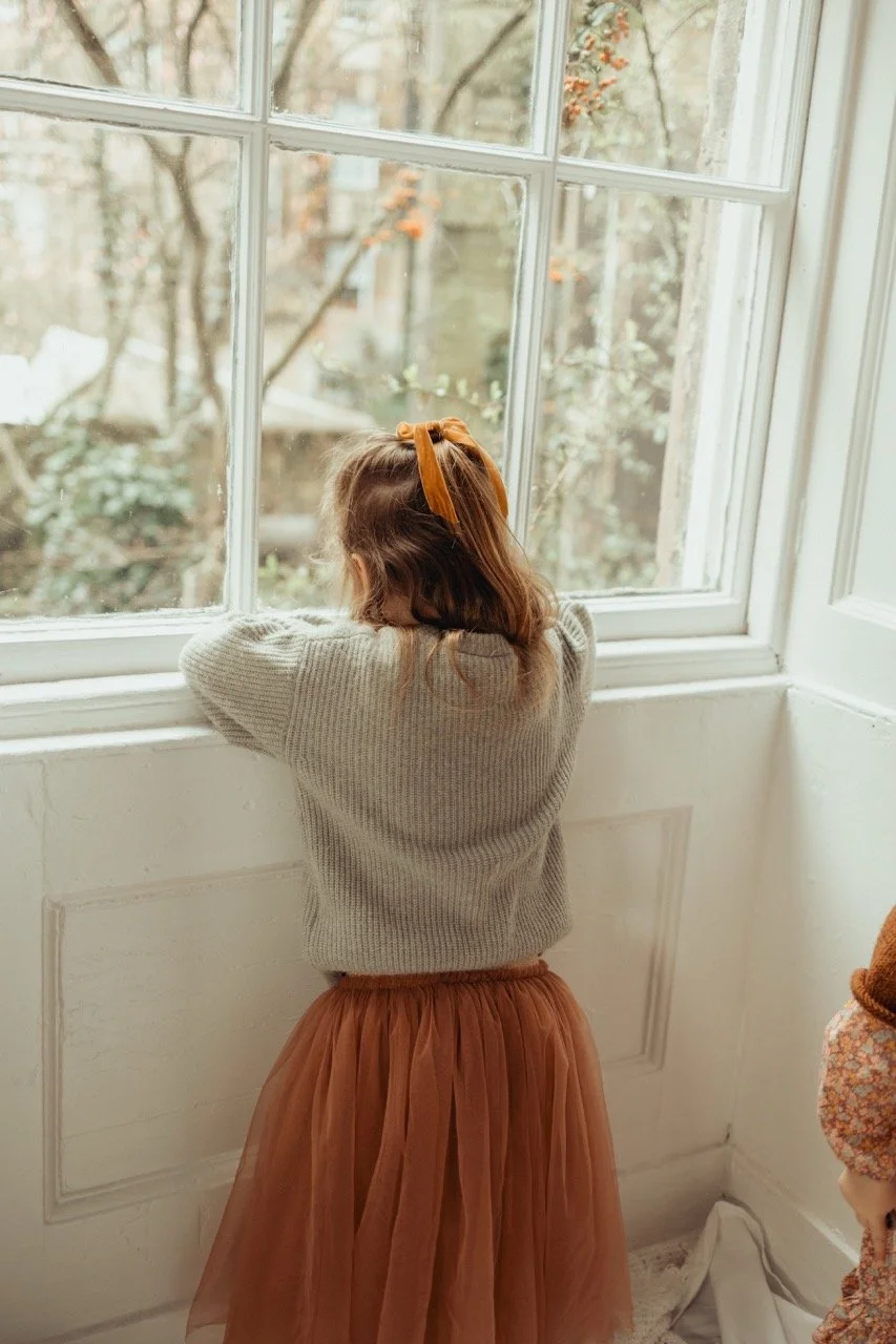 Child with a headband looking out a window wearing a sweater and skirt, room interior.