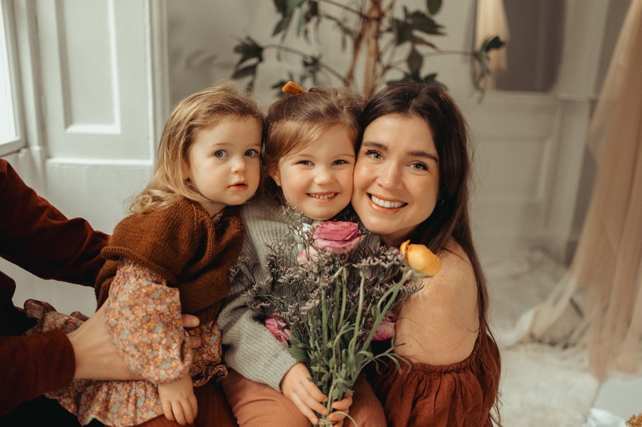 A woman smiling with two young girls, holding a bouquet of flowers indoors.