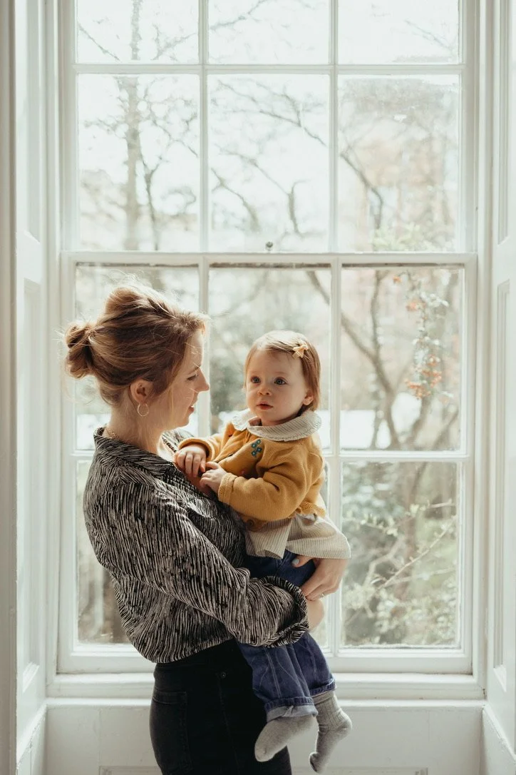 Woman holding a baby next to a large window with trees visible outside.