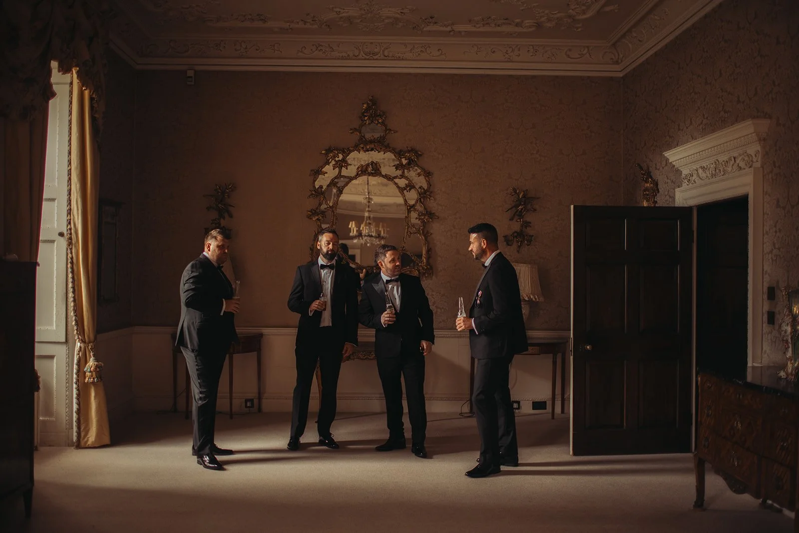 Four men dressed in tuxedos having a conversation and holding drinks in an elegant room with ornate decor, a large mirror, and a chandelier reflection.
