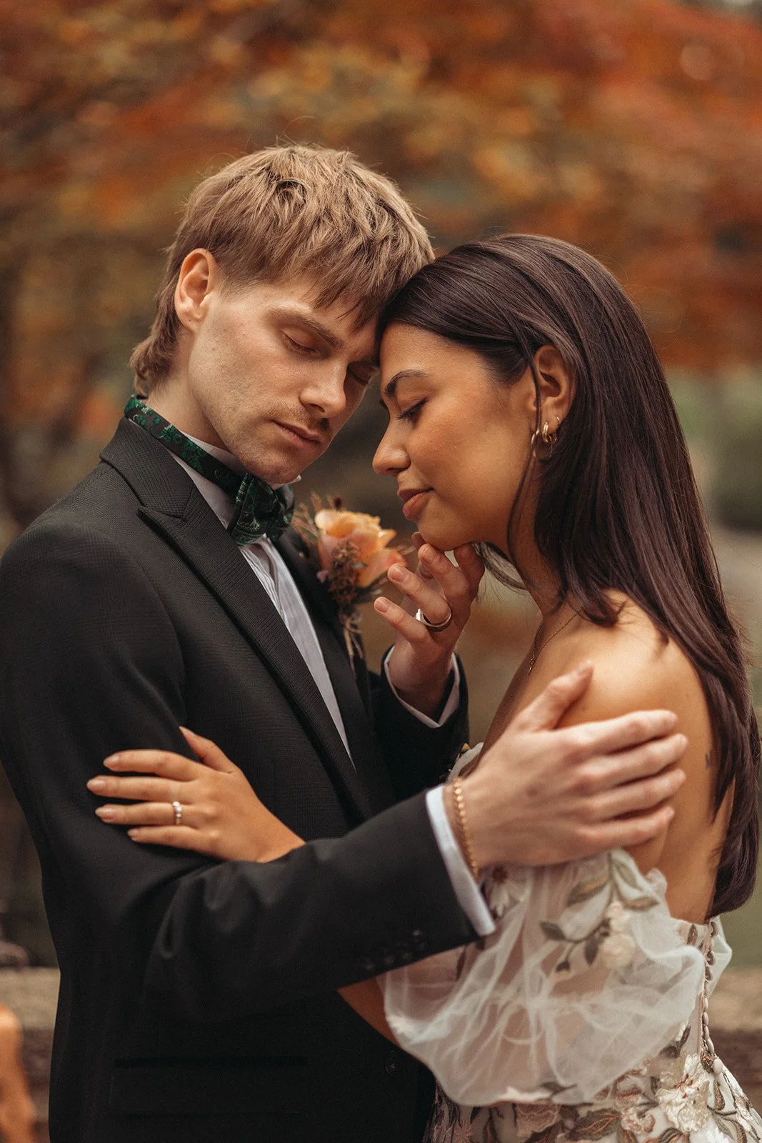 A man and woman embrace tenderly with foreheads touching, eyes closed, and a romantic setting with autumn foliage in the background.