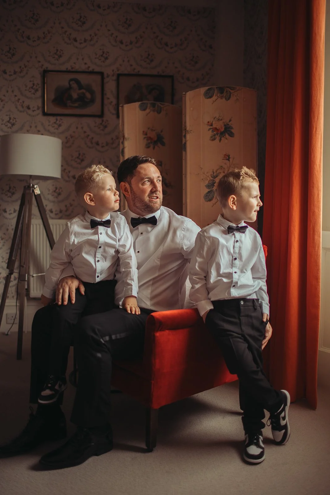 A man and two young boys dressed in white shirts with black bow ties, sitting and standing near a window with orange curtains. The man and boys are looking in the same direction outside the window. The room has floral wallpaper, a tripod lamp, and a 