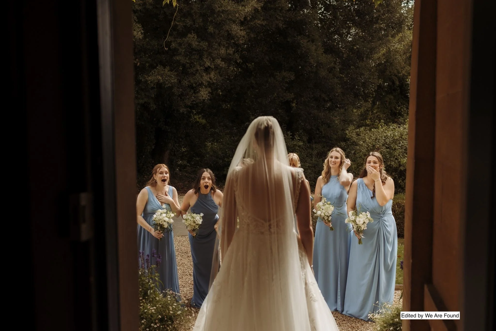 A bride wearing a white wedding gown and veil is seen from the back, facing a group of five bridesmaids in blue dresses, holding bouquets, and reacting excitedly outside during the daytime.