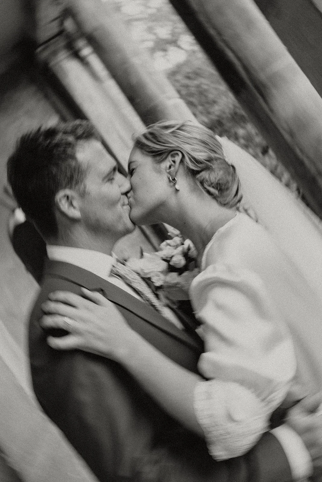 Black and white photo of a couple sharing a kiss on their wedding day, with the woman holding a bouquet of flowers and the man dressed in a suit and tie, standing outdoors near columns.