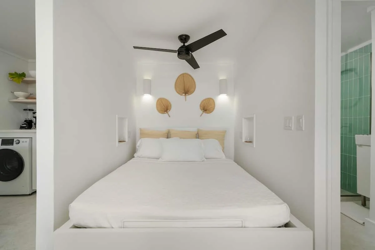 A minimalist bedroom with white walls and bed, decorative wall fans above the bed, small wall niches, and a ceiling fan. To the right, a bathroom with green tiles is partially visible.