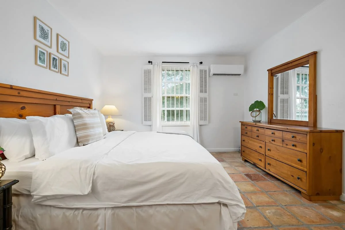 A bedroom with a wooden bed, white bedding, pillows, a nightstand with a lamp, a dresser with a mirror, and a window with white curtains.