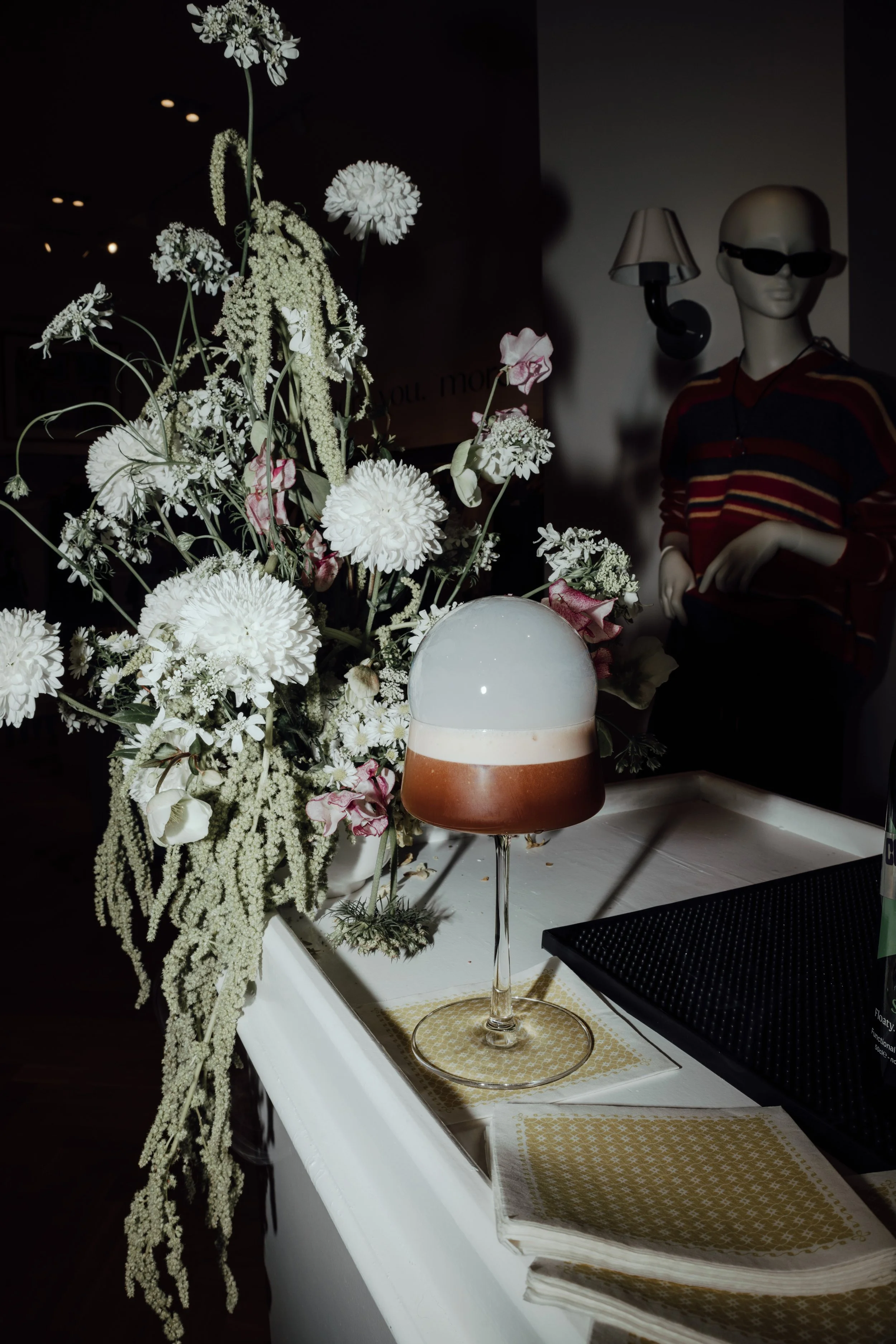 Zero-proof cocktail at Umbrella Dry Bar in Raleigh. A cocktail with two layers, one white bubble on top and one brown, in a coupe glass, placed on a table next to a bouquet of white and pink flowers.
