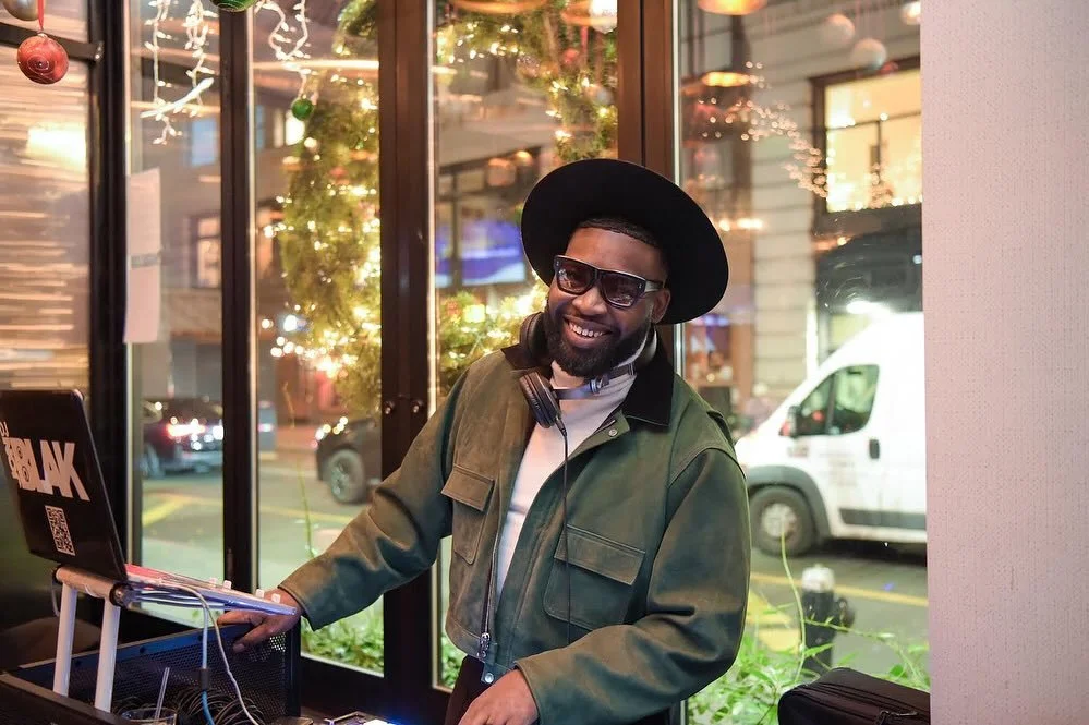 A smiling man with sunglasses and a black hat, wearing a green jacket with headphones around his neck, standing next to a DJ setup with a laptop, inside a decorated venue with Christmas lights outside the window.