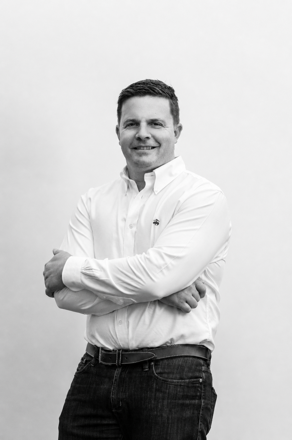 Black and white portrait of a smiling man with short dark hair, wearing a white button-down shirt and dark jeans, standing against a plain white background with his arms crossed.
