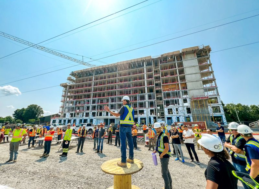 Construction site with a high-rise building under construction and a crowd of construction workers and supervisors wearing safety gear, gathered outdoors under a clear blue sky.
