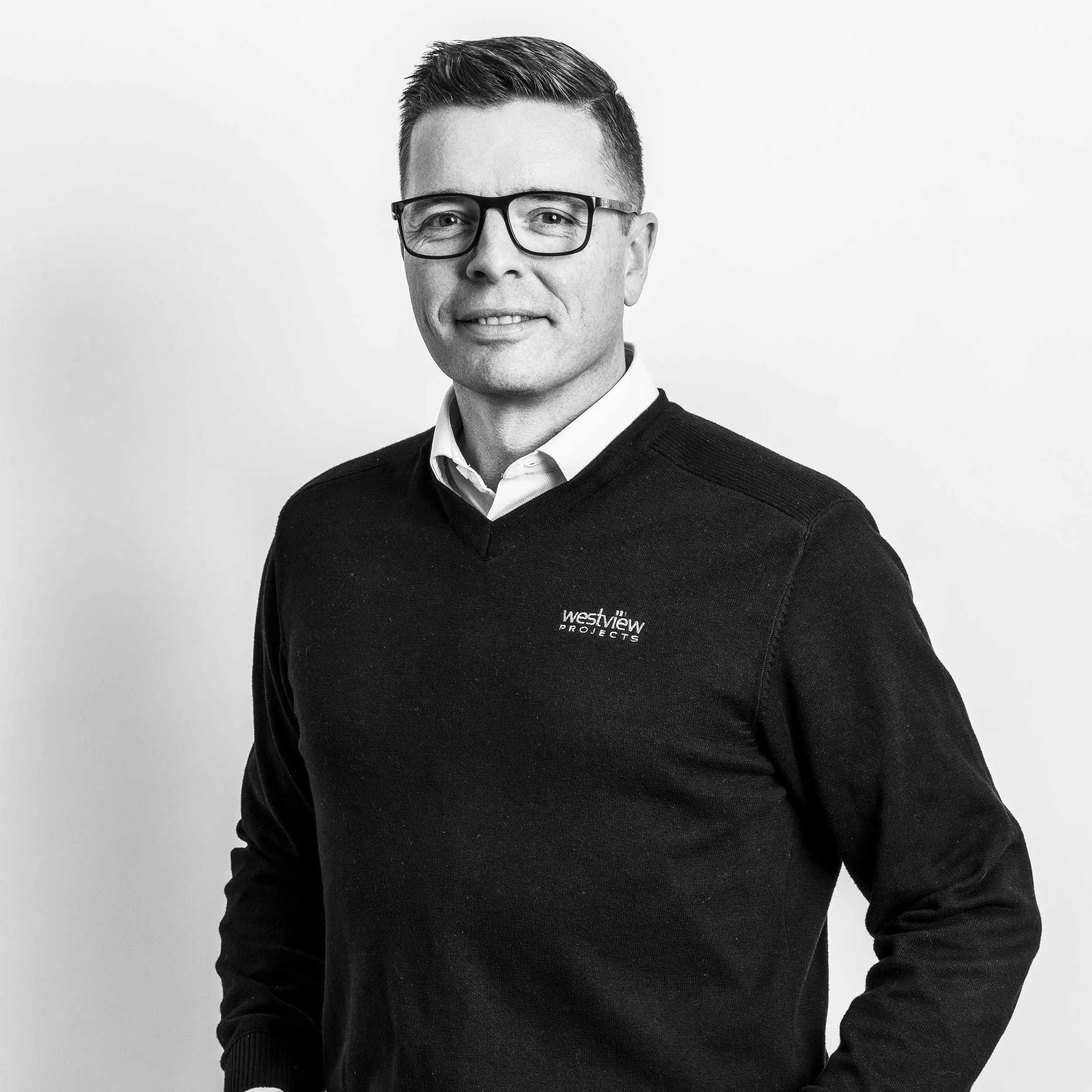 Black and white portrait of a man with short dark hair, wearing glasses, a white collared shirt, and a black sweater with 'Westview Projects' on it, standing against a plain white background.