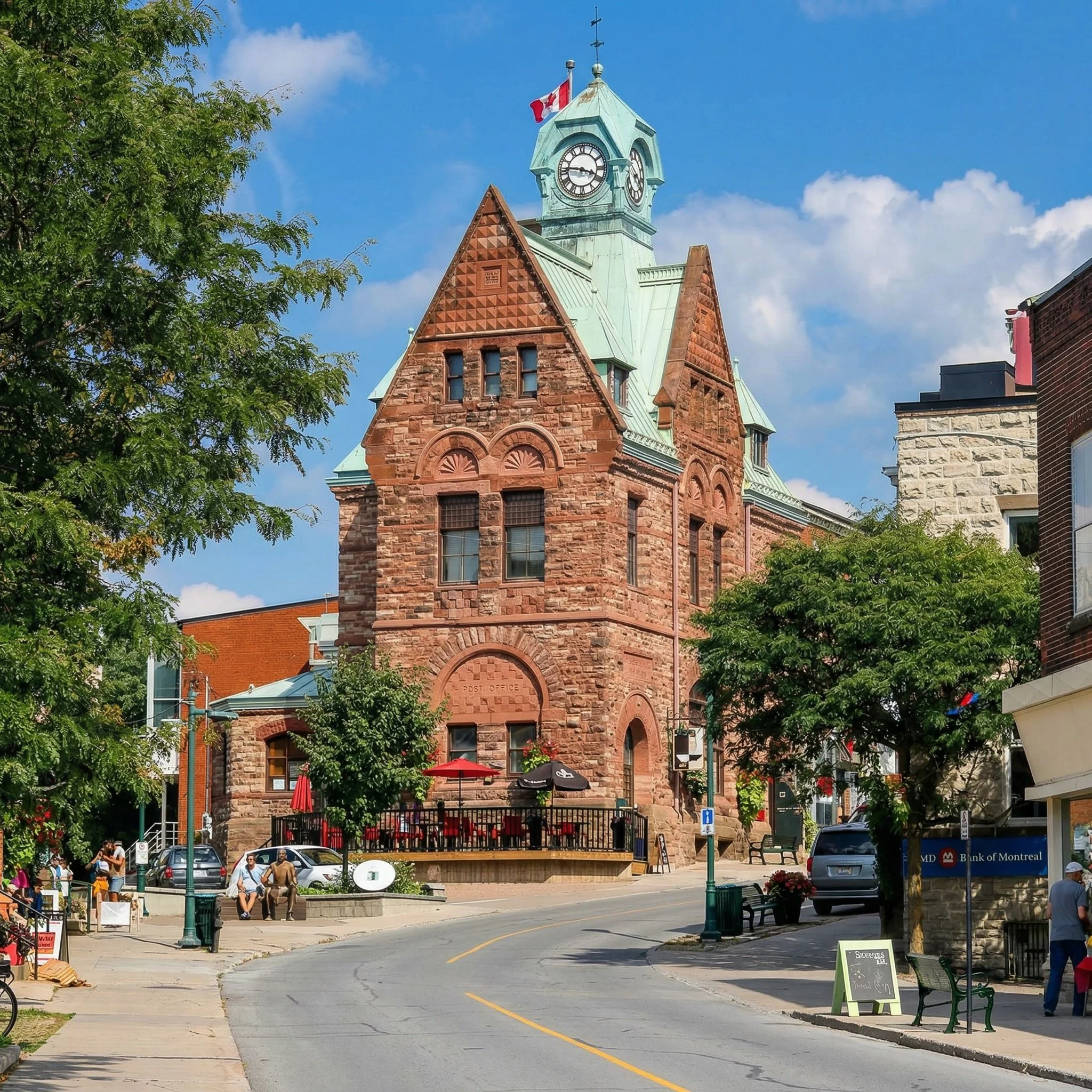 A historic red brick building with a clock tower and green roof, partially obscured by green trees, with people walking and sitting at outdoor tables in front, under a partly cloudy blue sky.