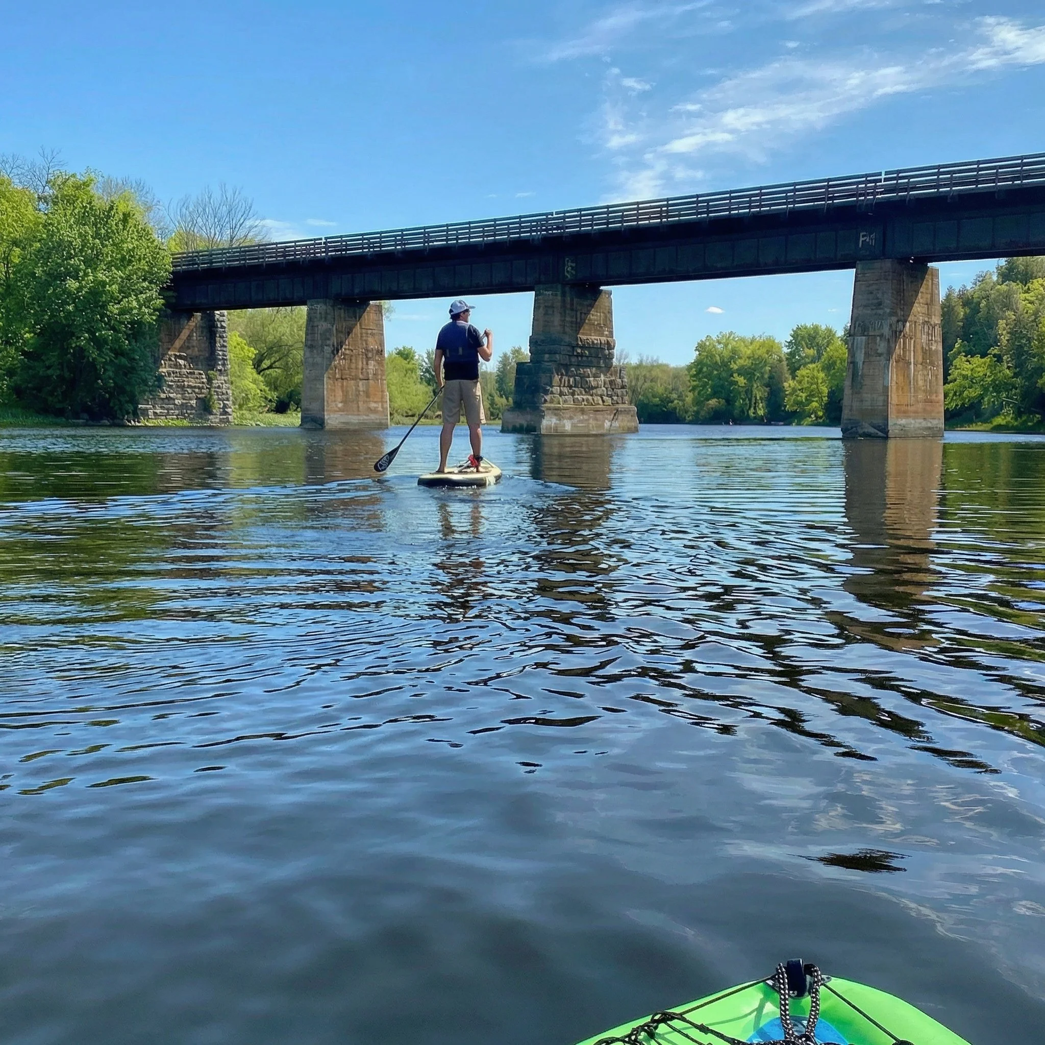A person paddleboarding on a body of water under a clear blue sky with a bridge in the background and green trees on the shore.
