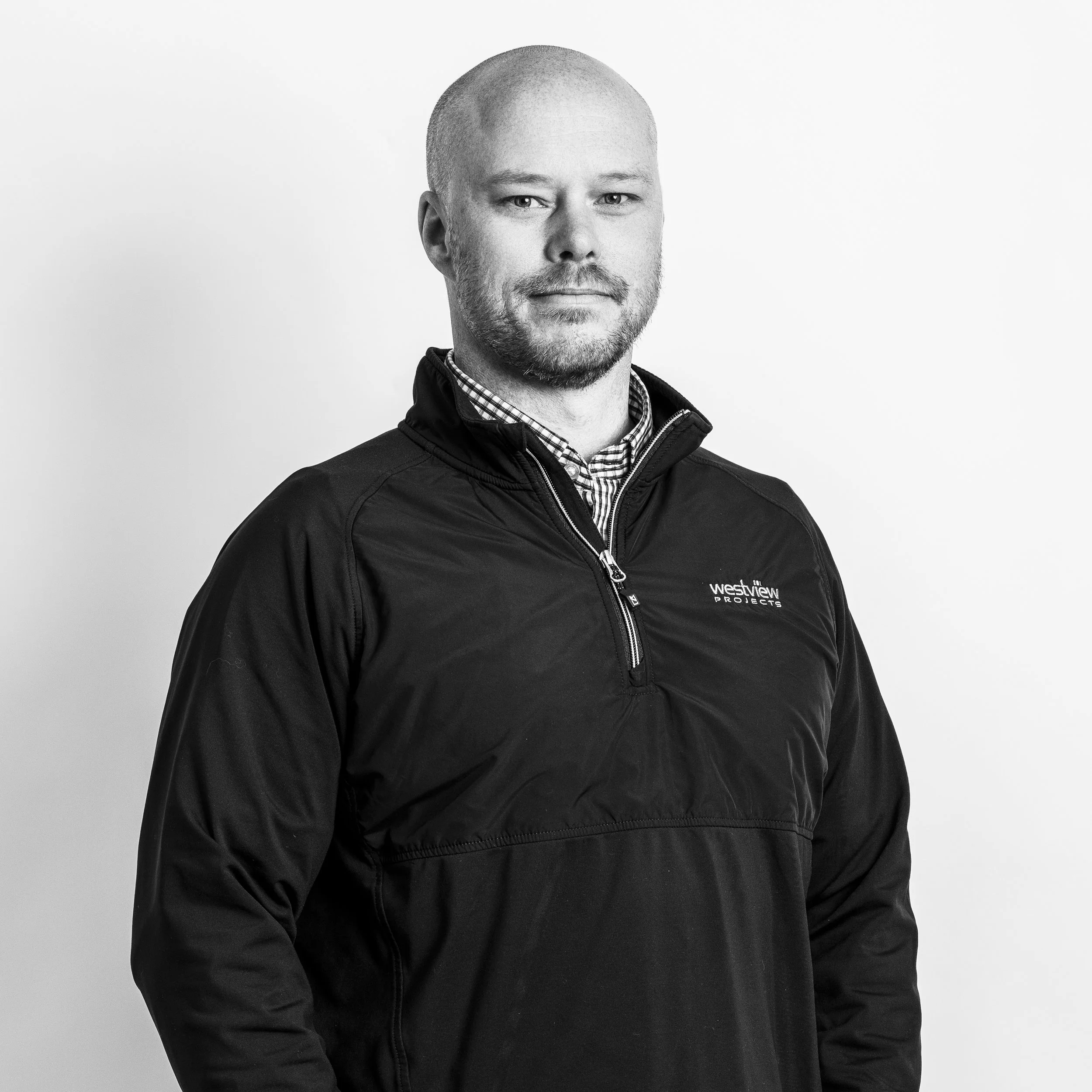 A man with a shaved head and facial hair wearing a black Westview Projects jacket and a checkered shirt underneath, standing against a plain white background.