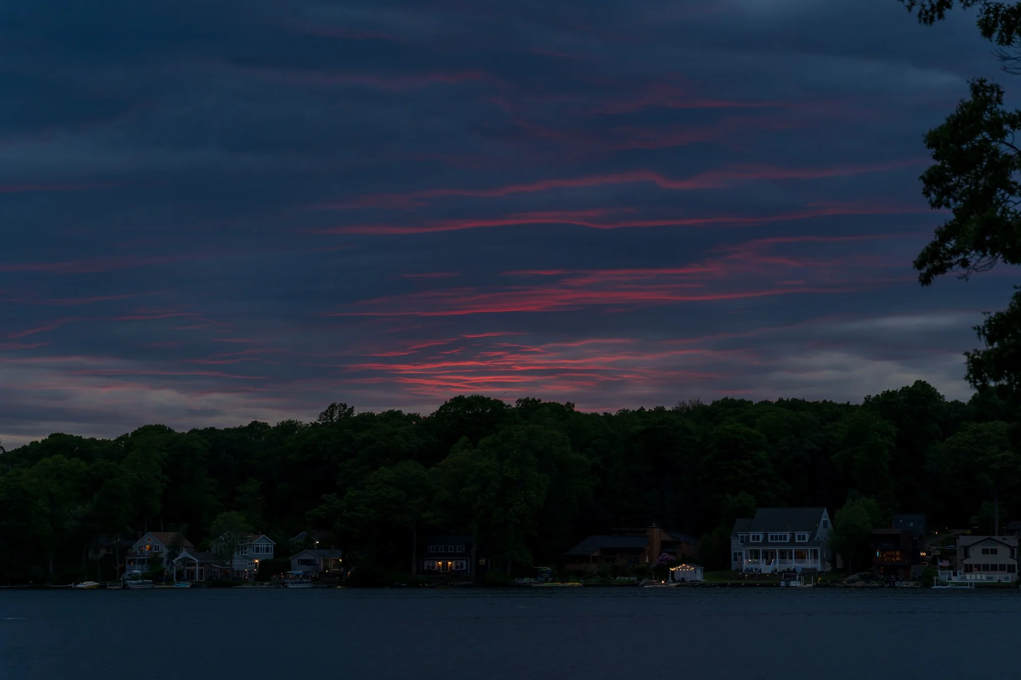 Dusk over a small lakeside community with houses and boats, dark clouds with pink and purple hues in the sky, and trees silhouetted against the horizon. Located on Lake Pocotopaug in East Hampton, CT.