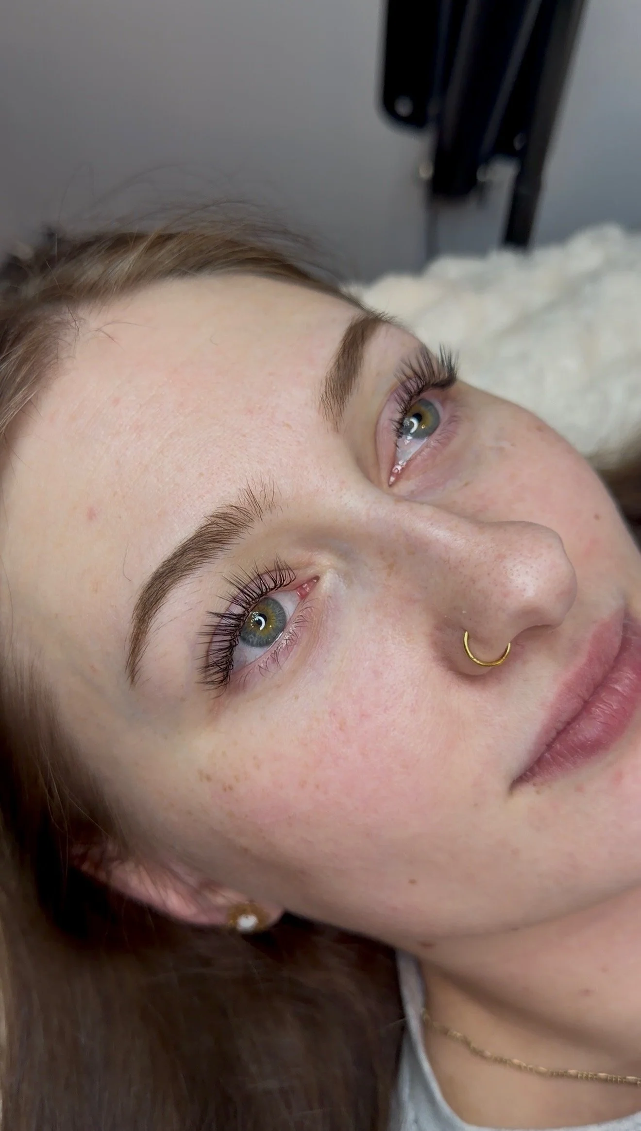 Close-up of a woman lying down with her eyes open, showing her face from her forehead to her chin, with long eyelashes, light freckles, and a gold nose ring.