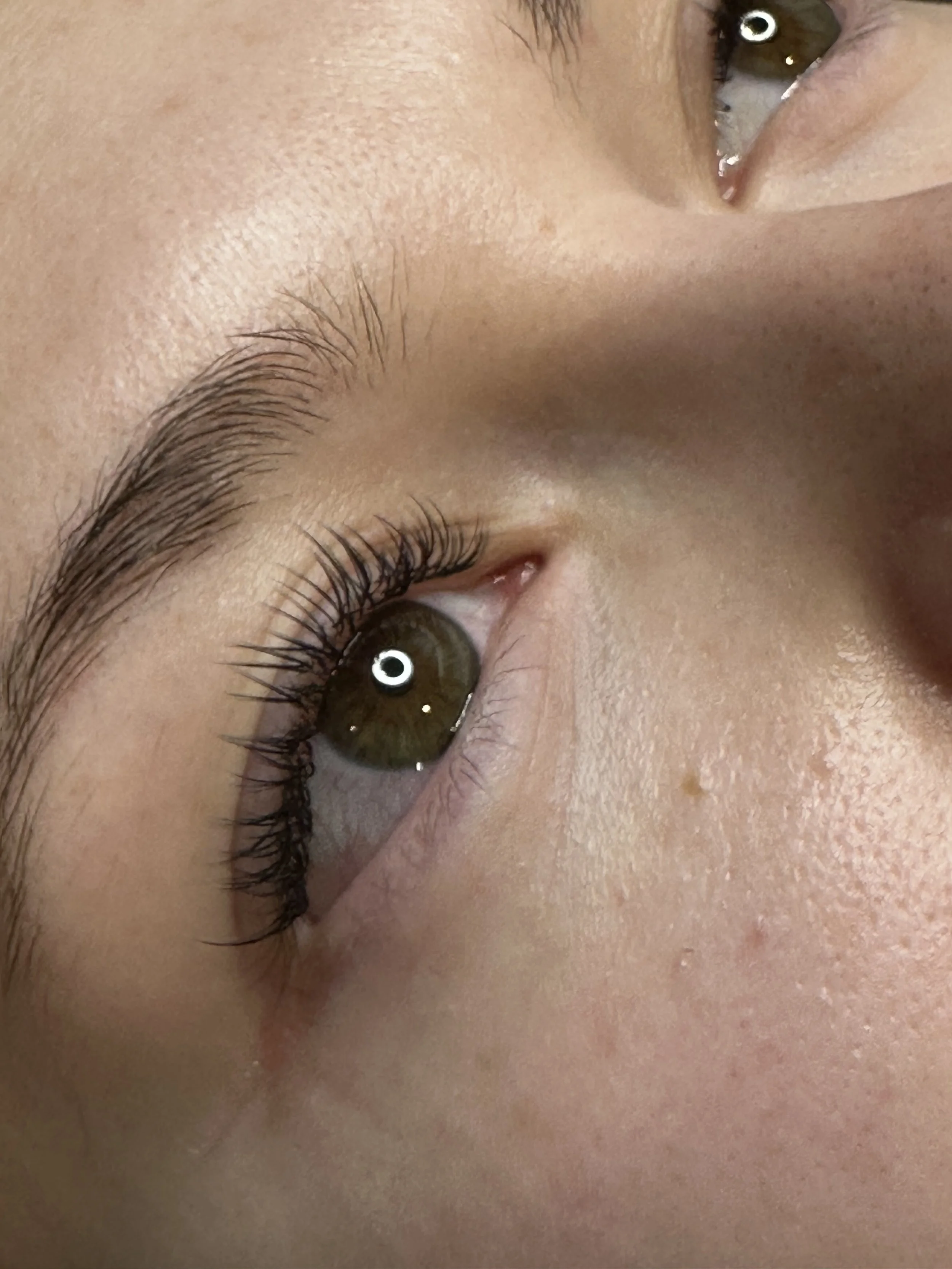 Close-up of a person's eye, showing the eyelash, eyebrow, and part of the nose. The eye has a greenish-brown iris and reflects light.