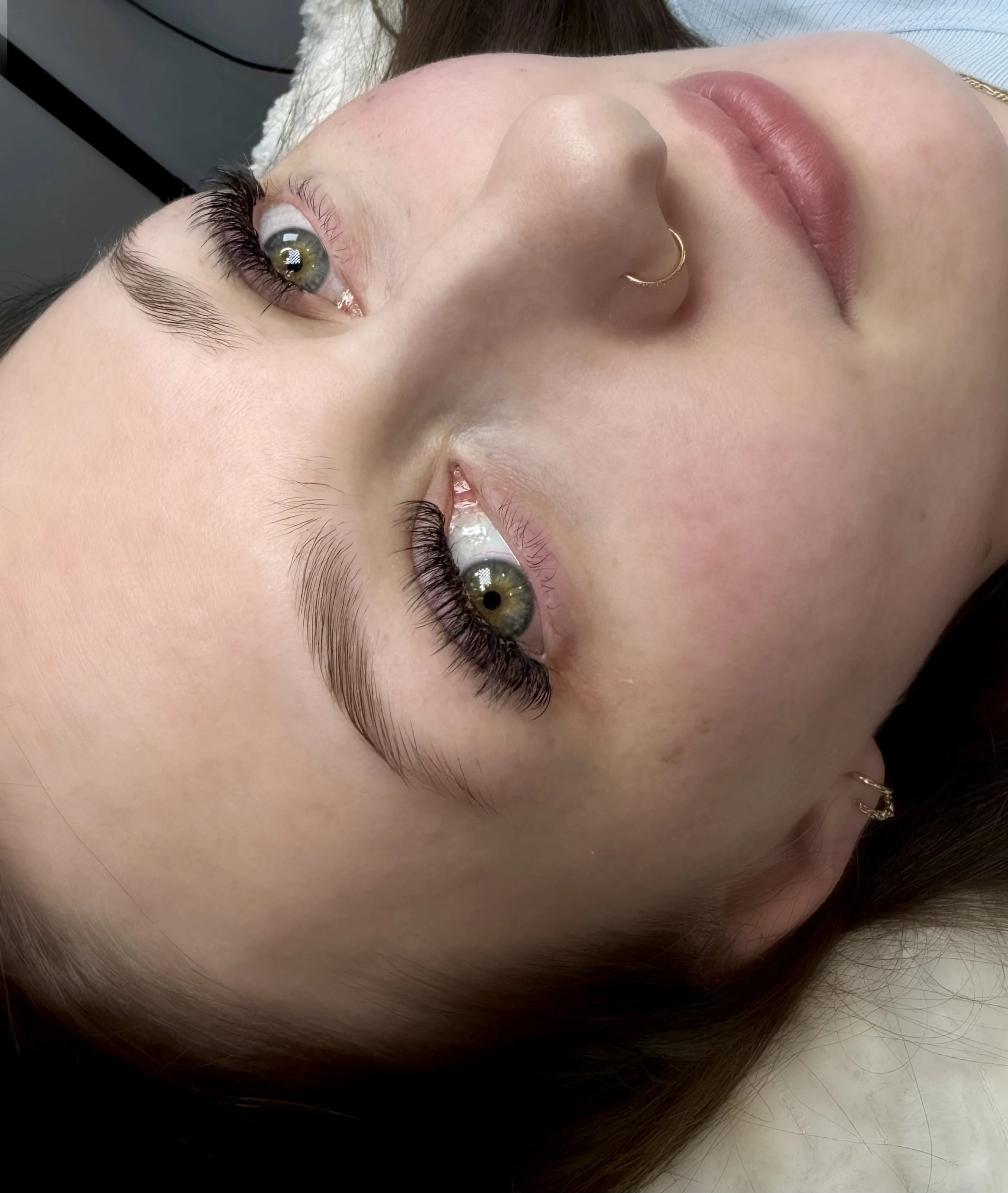 Close-up of a woman's face lying down, showing her green eyes, long eyelashes, and nose with a gold hoop piercing. Her lips are slightly parted.
