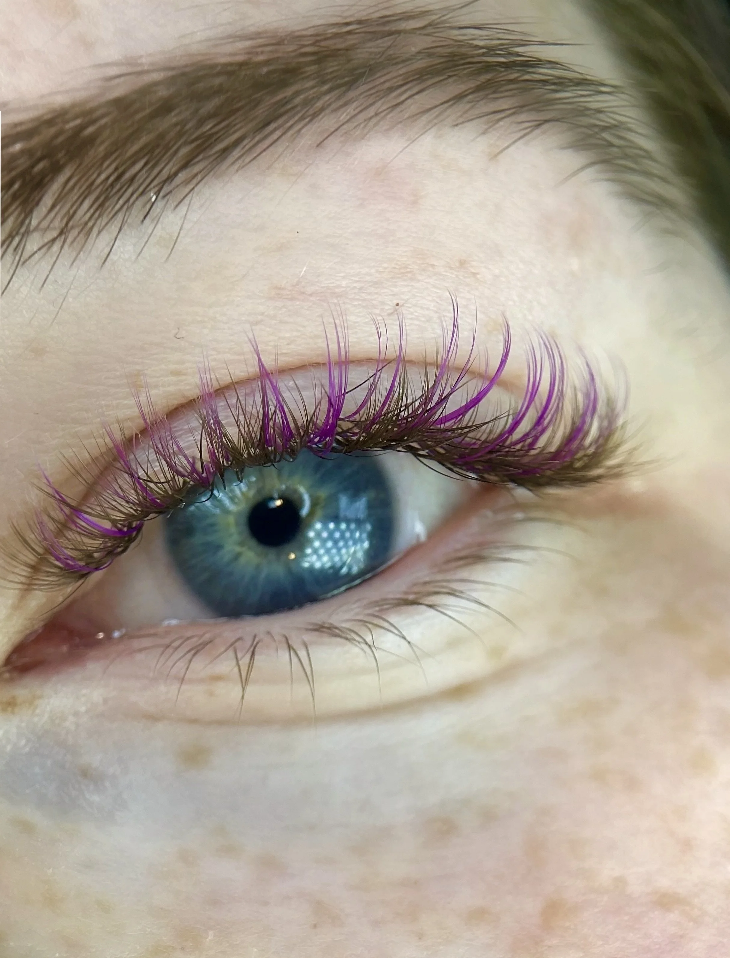 Close-up of a person's eye with blue iris, long eyelashes with purple and black mascara, and a natural eyebrow.