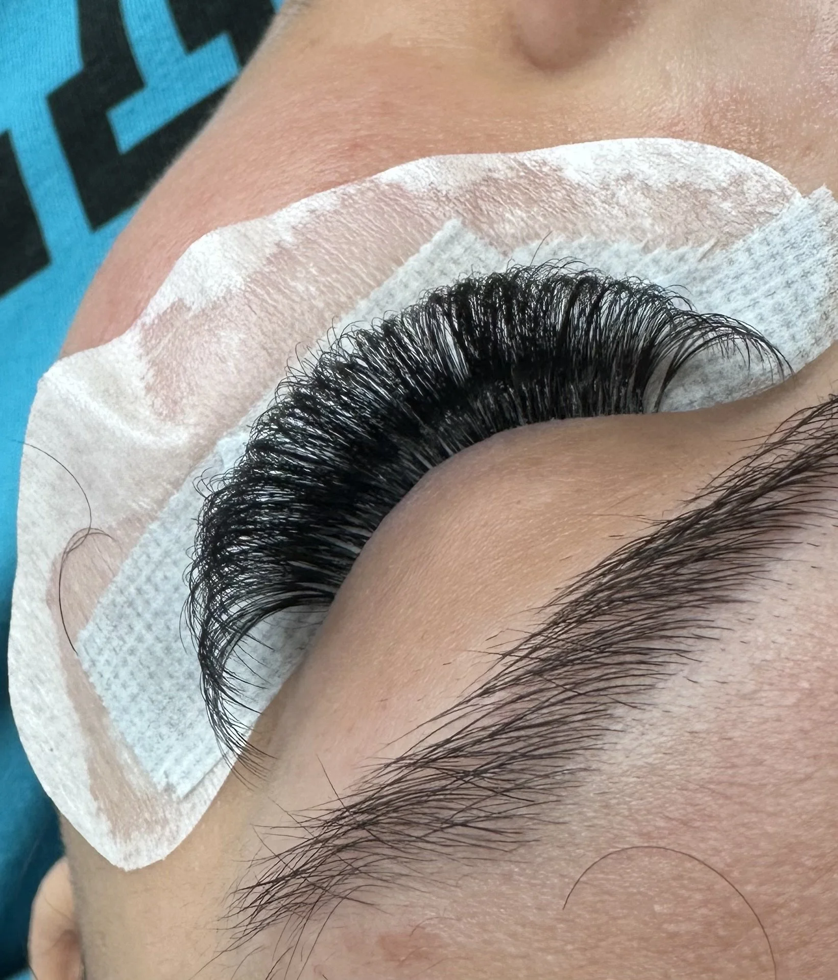 Close-up of an eye with long black eyelashes resting on a skin tone, partially covered by a white adhesive eyelash extension pad.