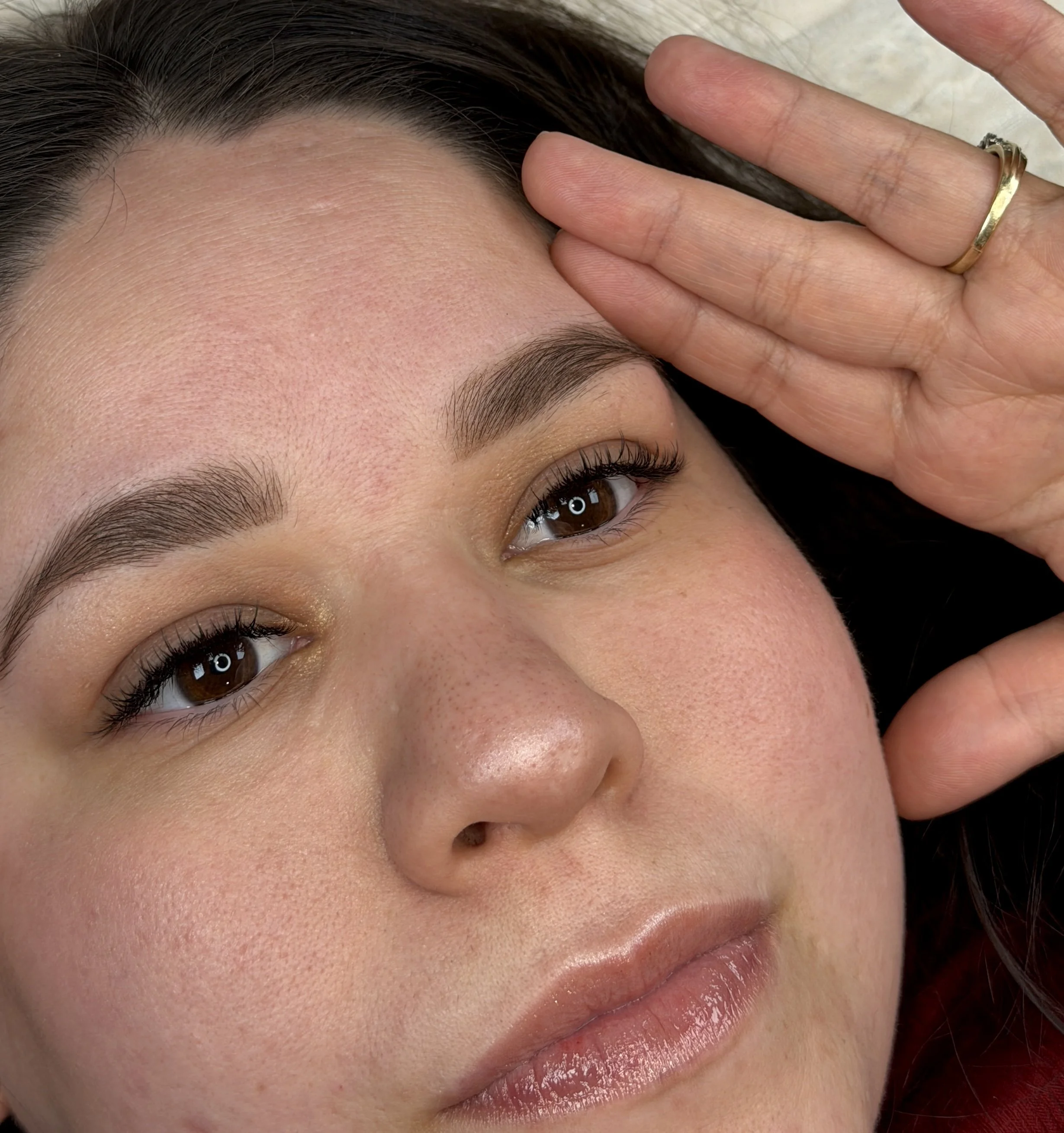 Close-up of a woman's face showing her eye, eyebrow, forehead, and lips. She is resting her head on a surface and has her hand with a gold ring on her finger near her forehead.