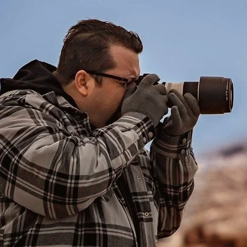 A man in a plaid jacket and glasses takes a photograph with a camera outdoors.