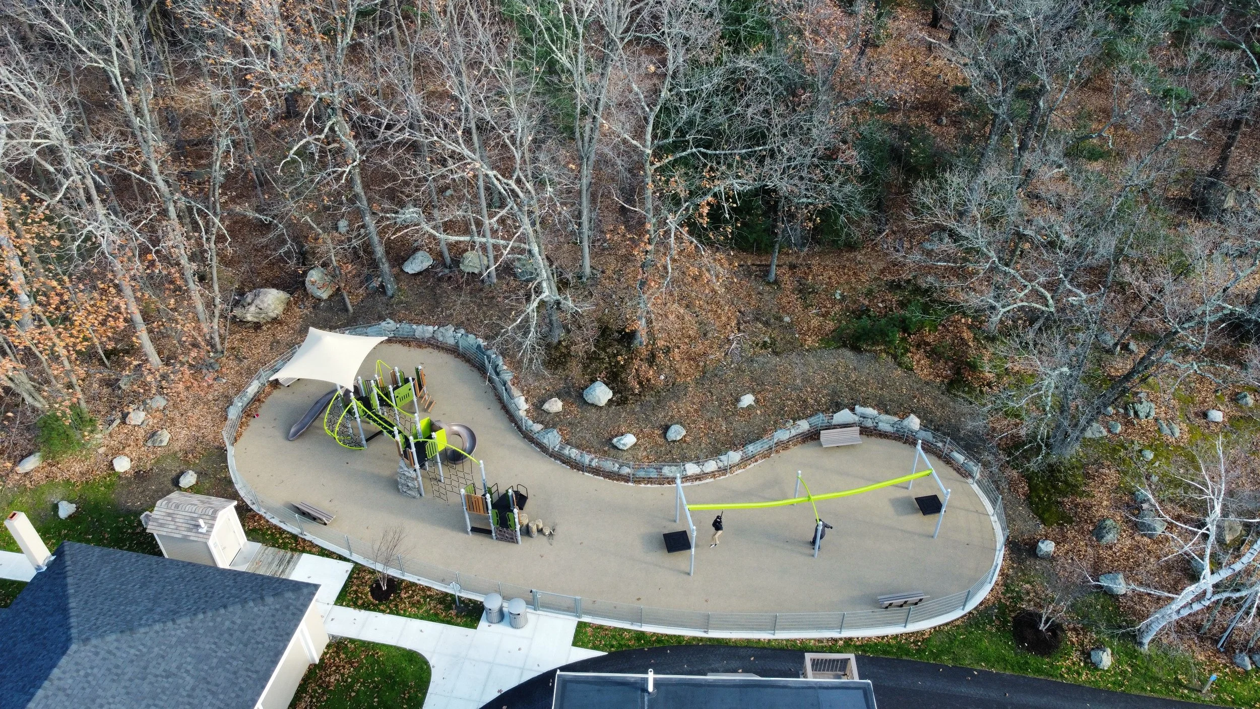 Aerial view of a playground with green play structures and a canopy, surrounded by bare trees and fallen leaves.