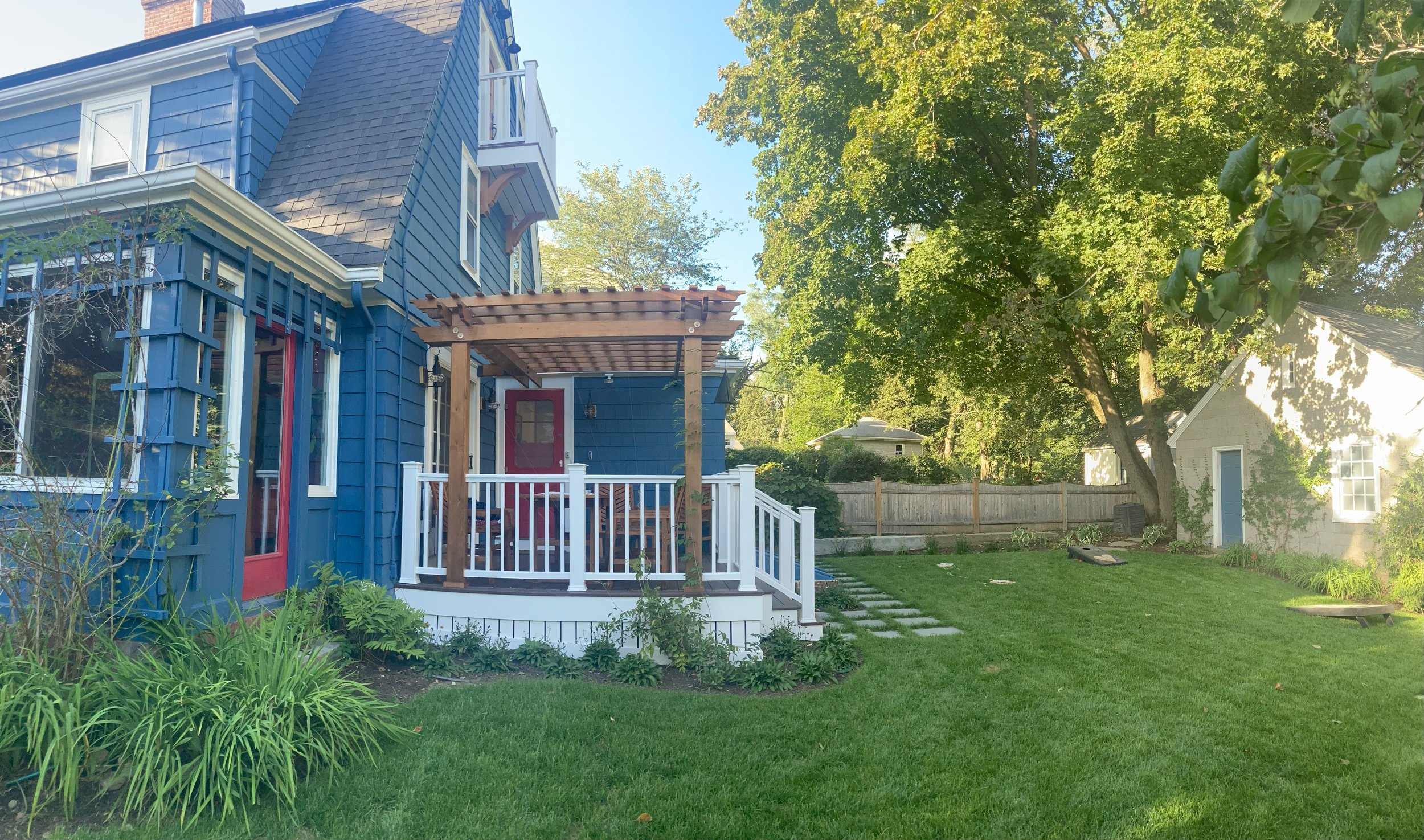 Blue house with red door, covered porch with pergola, lush green lawn, and large tree in backyard, bordered by a wooden fence.
