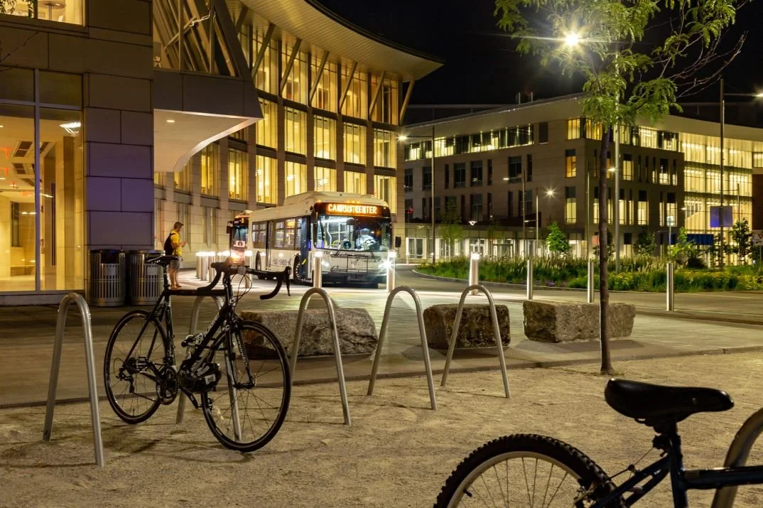 Night scene at a university campus with a bus labeled "Campus Center," bicycles parked at a rack, a modern building with illuminated windows, and green landscaping.