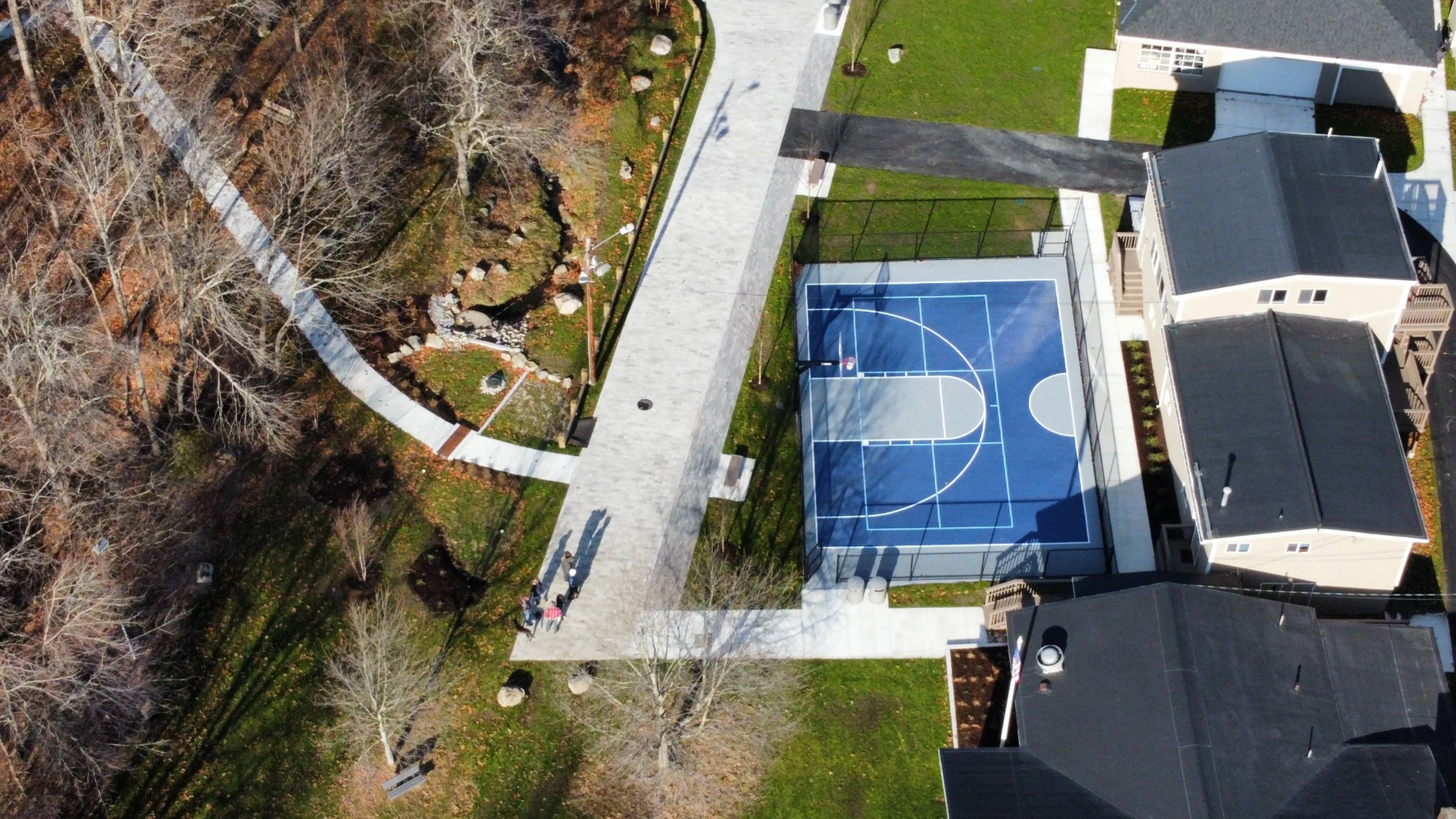 Aerial view of a residential area featuring a blue outdoor basketball court, adjacent pathways, several houses with dark roofs, trees, and landscaped greenery.