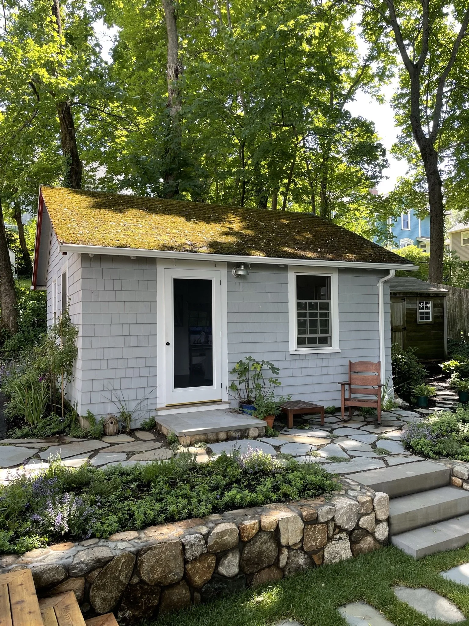 Small gray cottage with moss-covered roof, surrounded by lush green trees and a stone-paved patio with plants and a wooden chair.
