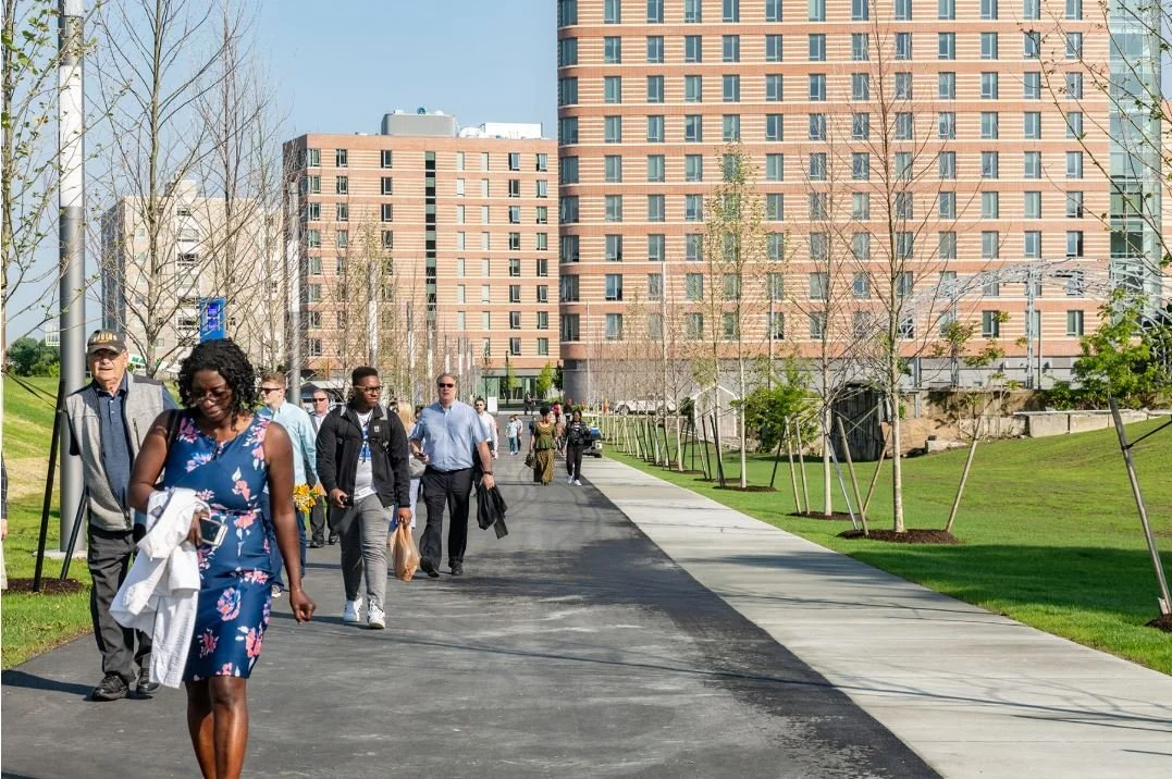 People walking on a path lined with trees and greenery, with tall brick buildings in the background.