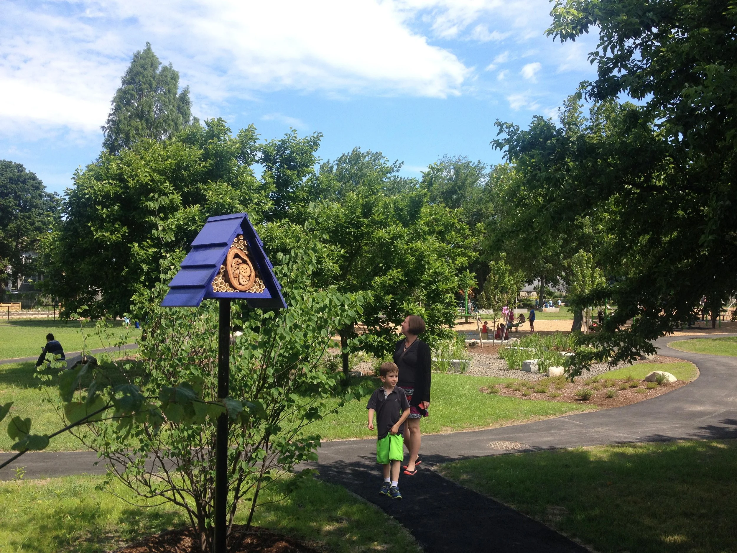 Park scene featuring a winding path, trees, and a blue birdhouse. People in the background are walking and sitting. A child and an adult are in the foreground.