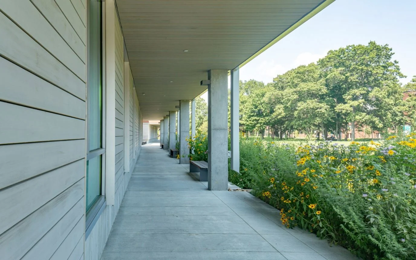 Covered walkway with concrete columns, wooden paneling, and a garden featuring yellow flowers to the right.